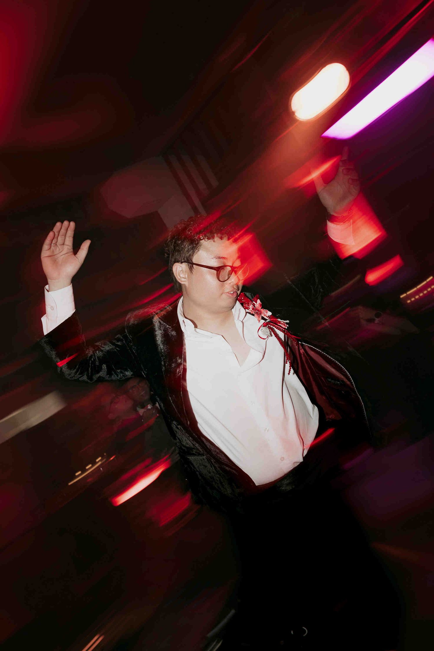 Man in black blazer and red glasses dancing energetically under red stage lights at a party.