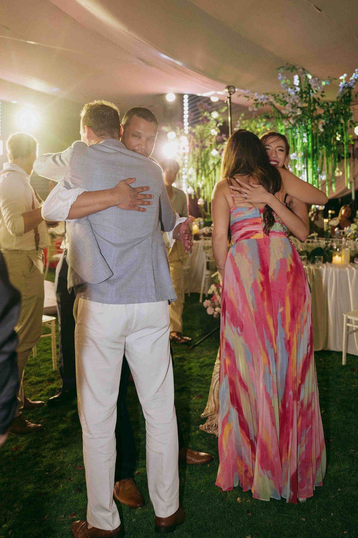 Couples embracing at an outdoor wedding reception under a tent with string lights and lush greenery.