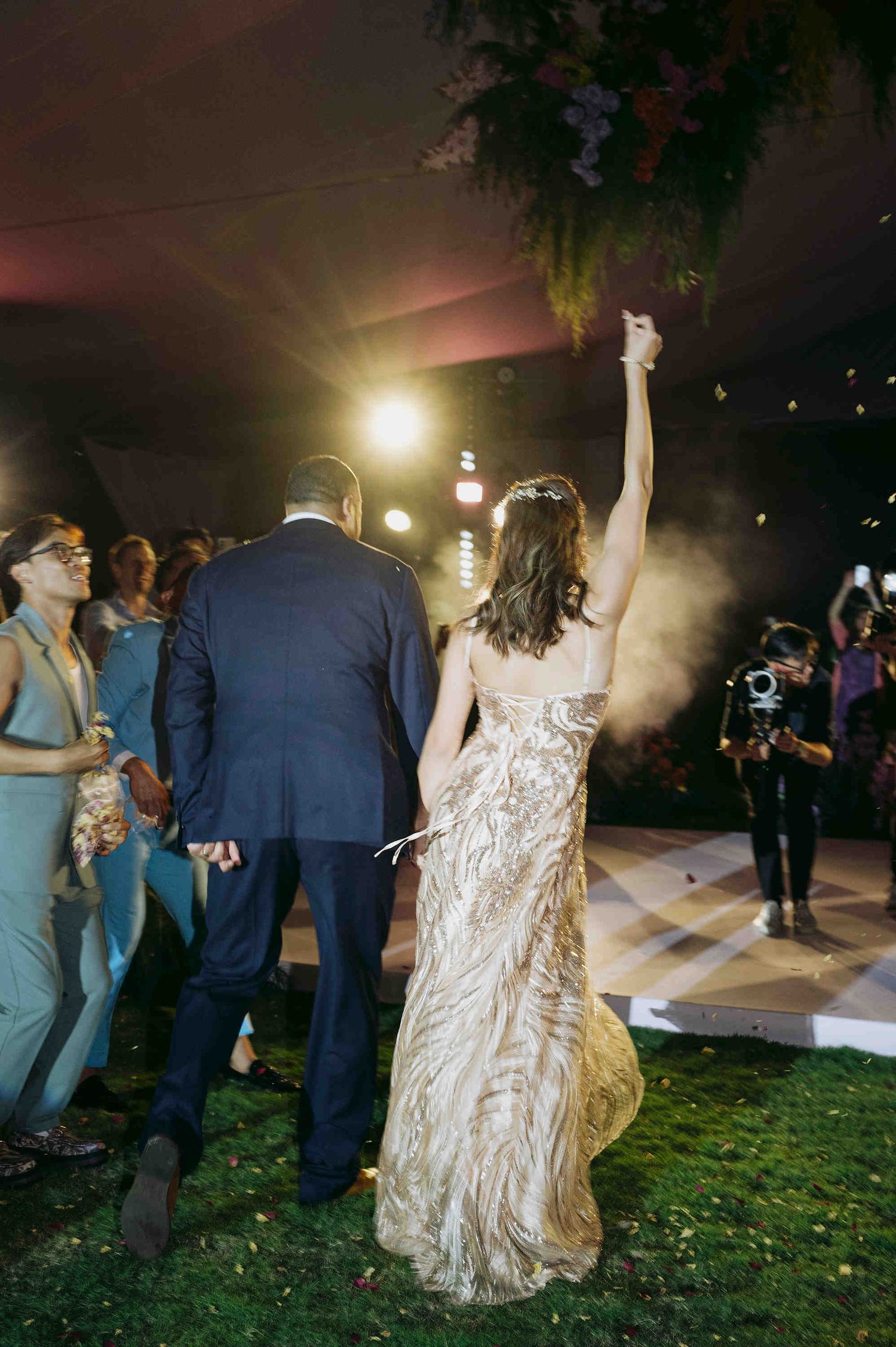 Bride in gold gown raises arm celebrating with groom in navy suit at outdoor wedding reception at night.