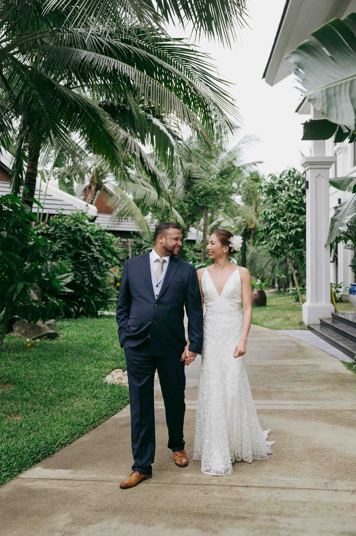 Bride and groom holding hands walking on tropical resort path, she in lace gown, he in navy suit.