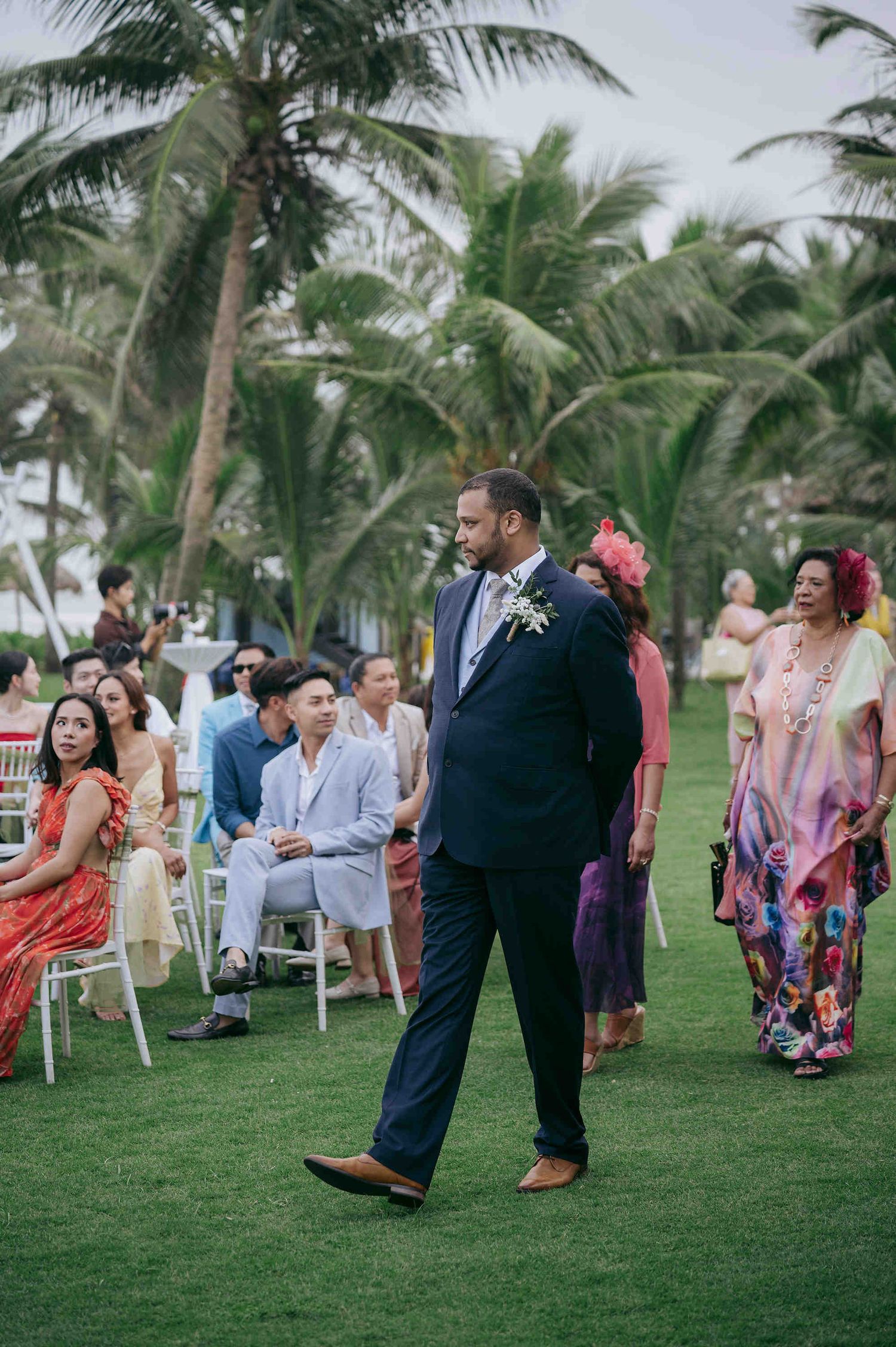 Groom in navy suit walks down outdoor wedding aisle flanked by guests in colorful attire, with tropical palm trees in background.