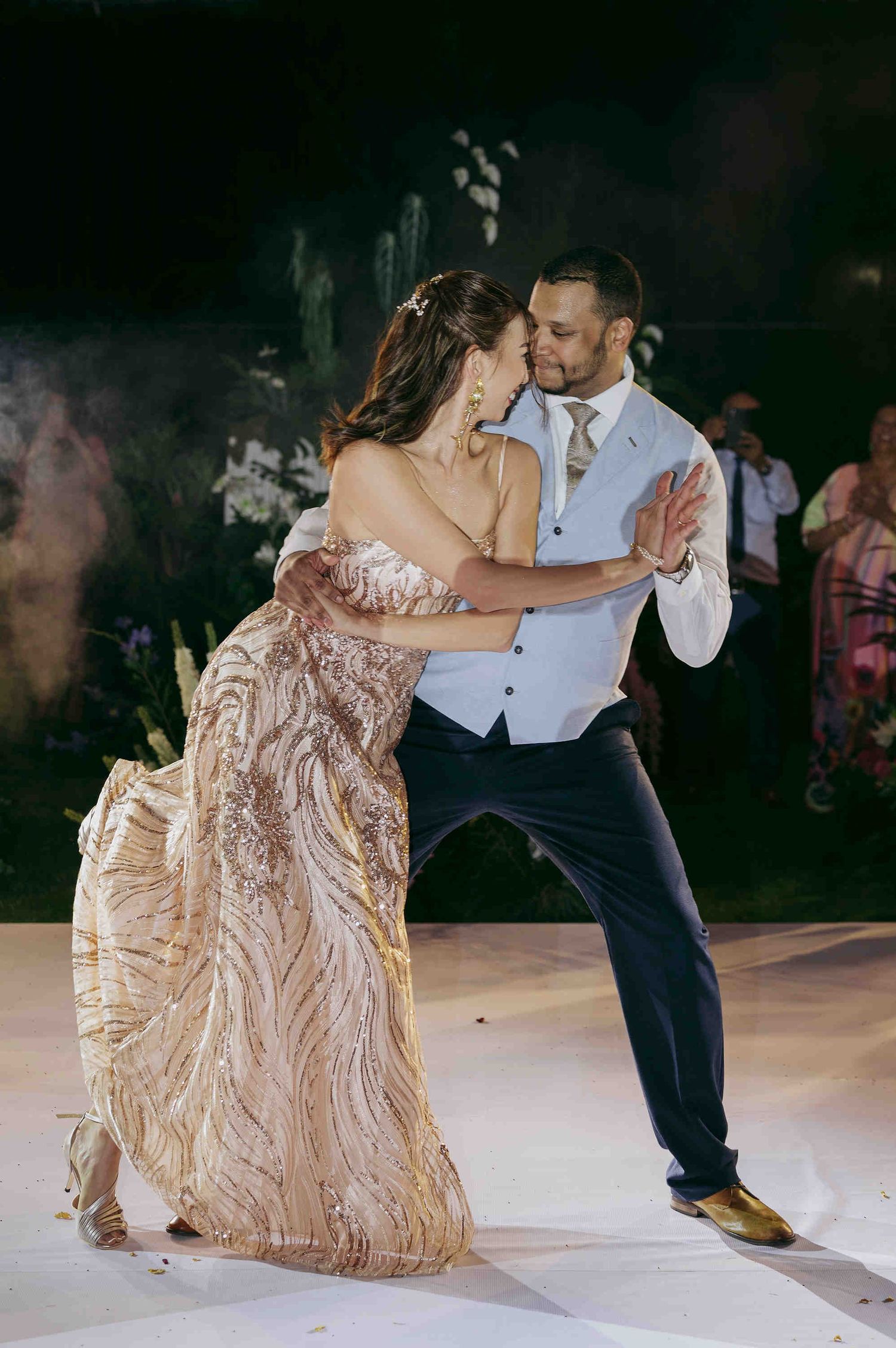 Couple sharing their first dance at wedding, bride in gold sequin gown, groom in light blue vest and navy pants.