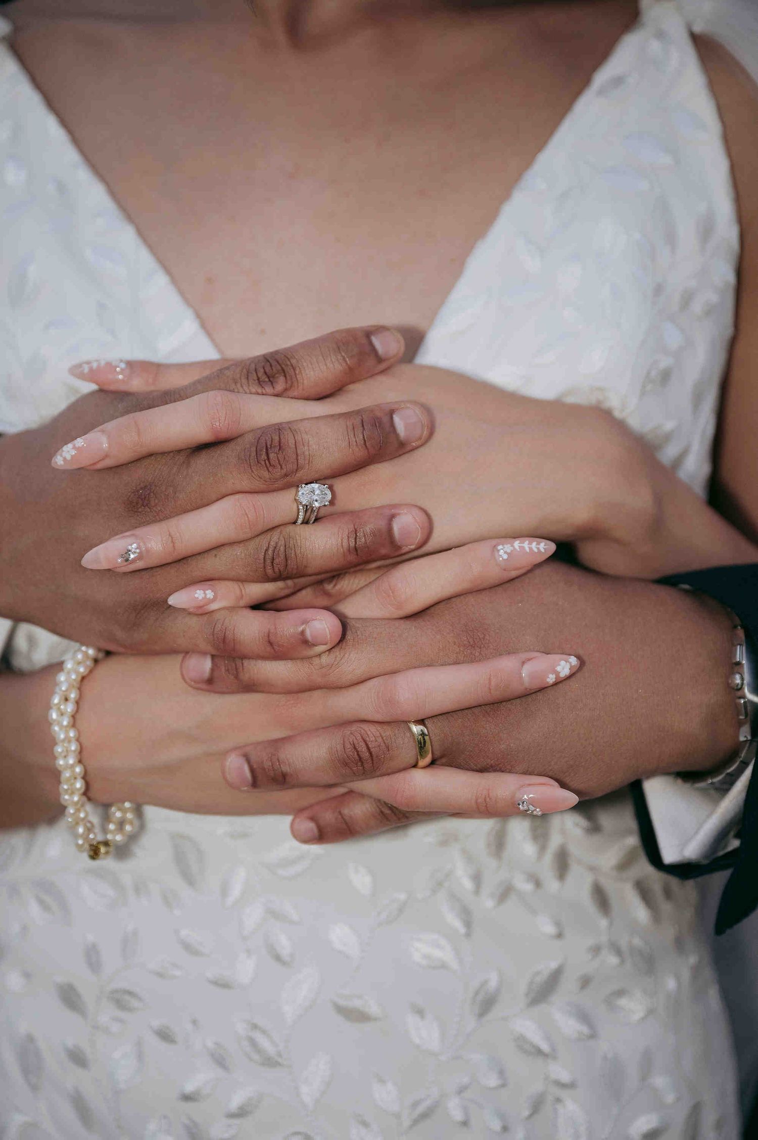 Close-up of newlywed couple's hands showing engagement ring, wedding bands, pearl bracelet, and elegant white nail art.