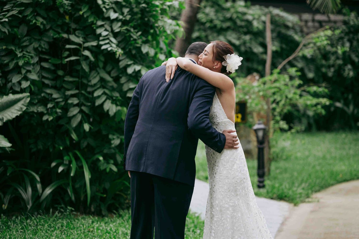 Bride in lace wedding gown embracing a man in a navy suit outdoors, surrounded by lush green tropical foliage.