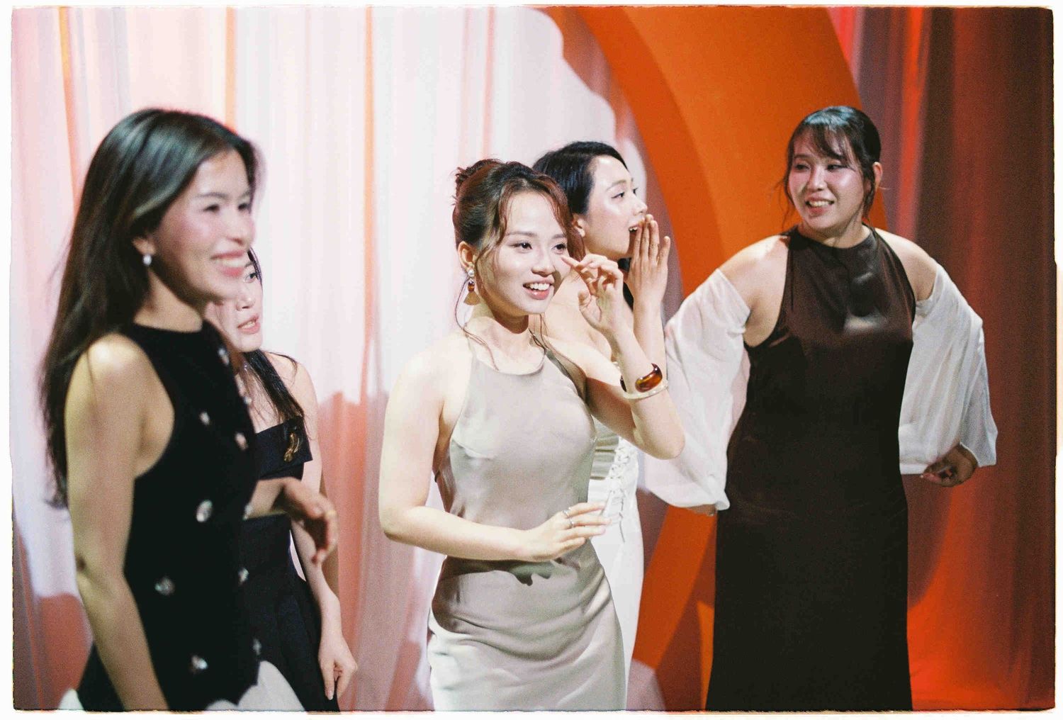 Four young Asian women in elegant dresses laugh and pose together on a colorful stage backdrop.