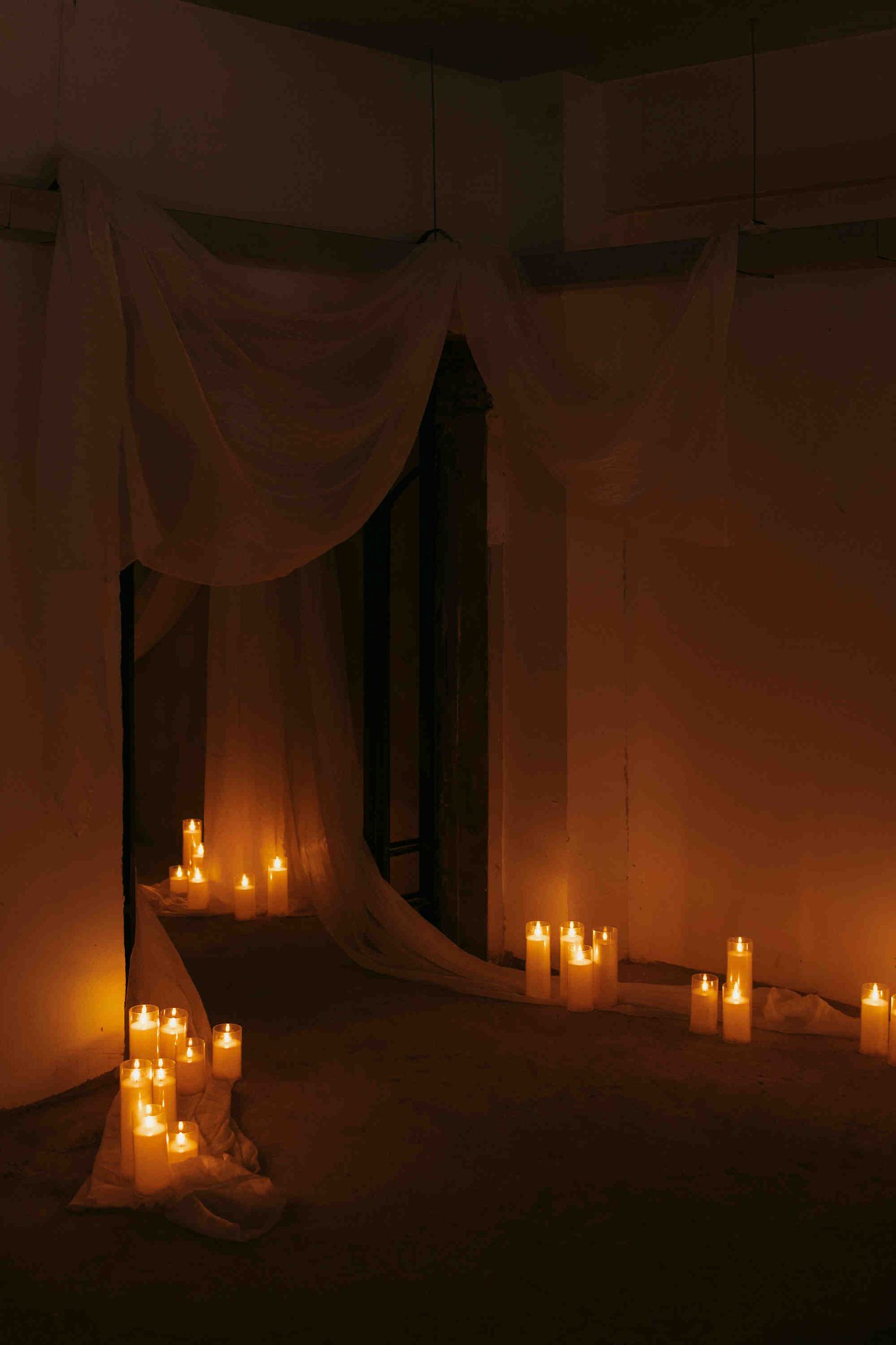 Romantic candlelit room with white draped fabric and glowing pillar candles on the floor creating a warm, intimate atmosphere.