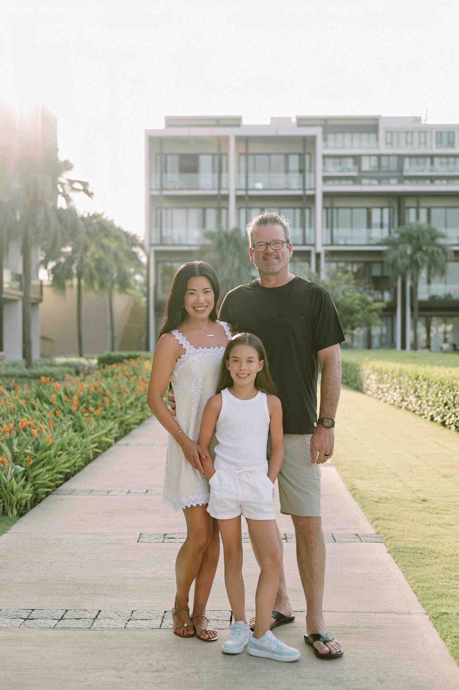 Da Nang family: family of three poses together on a sunny walkway with tropical landscaping and resort buildings visible, by AnhVuWedding.