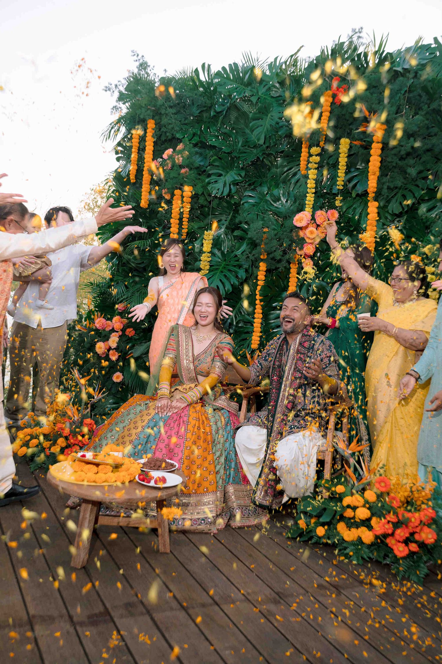 Couple in vibrant Indian wedding attire celebrate with flower petals and marigold garlands at a colorful outdoor ceremony.