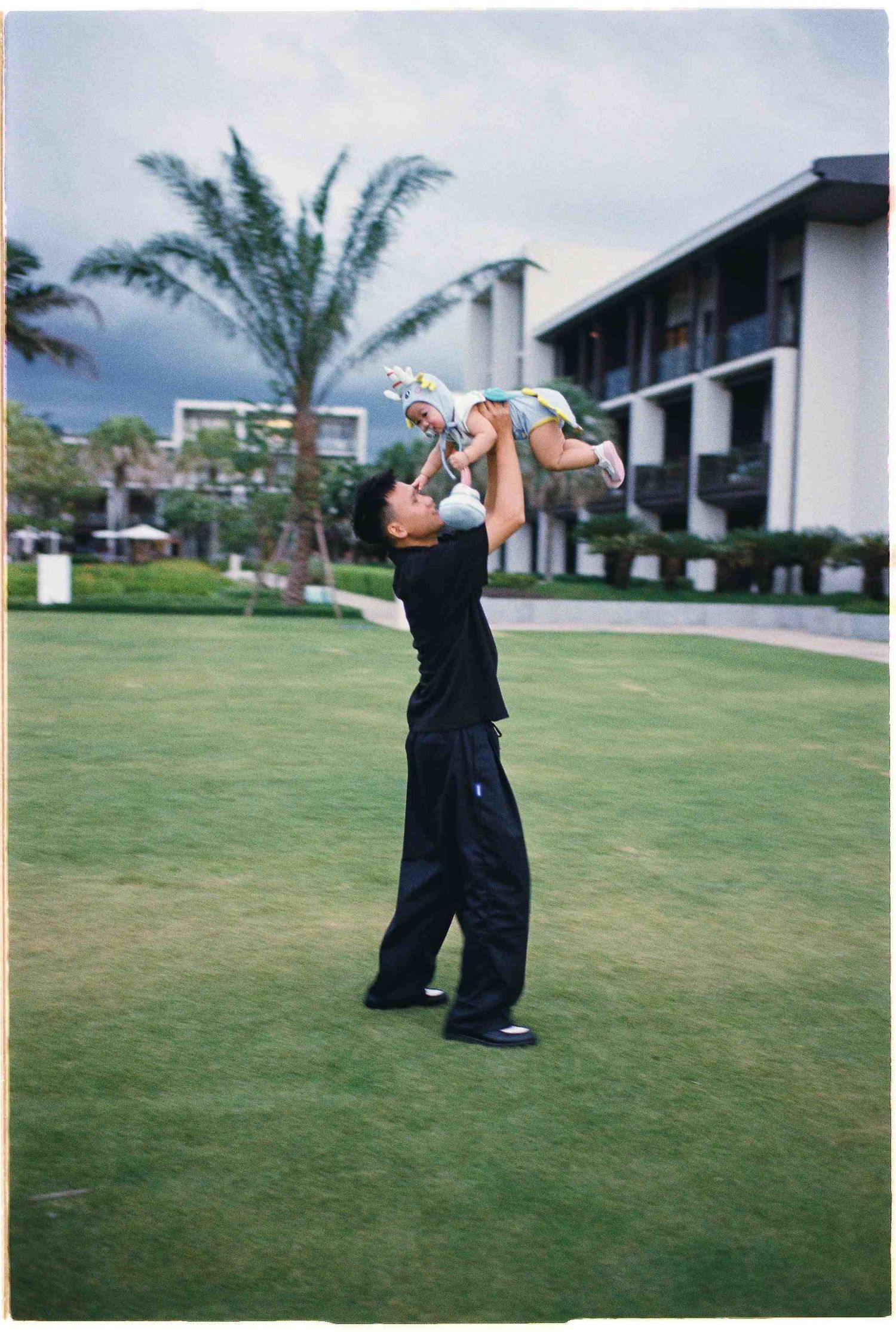 Da Nang family: adult lifts a baby high in the air while playing on a grassy lawn with resort buildings behind them, by AnhVuWedding.