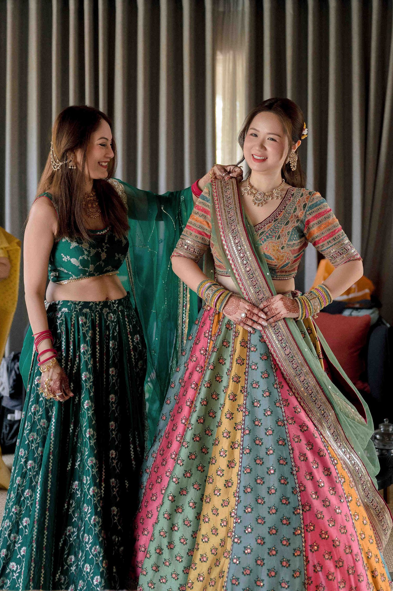 Two women smiling in vibrant Indian lehenga outfits, one multicolored and one dark green, adorned with traditional jewelry.