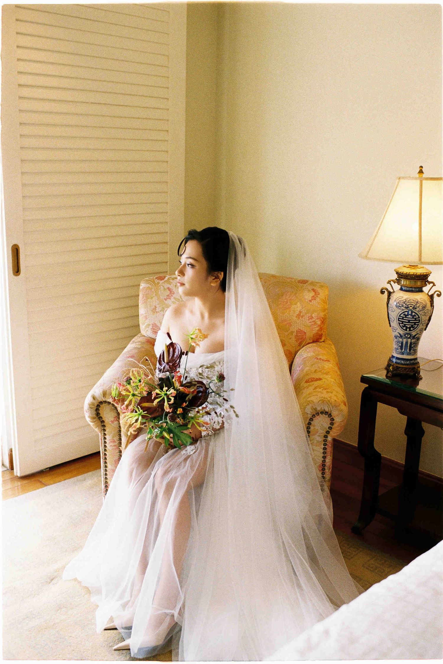 Bride in white gown and veil sits on gold chair holding floral bouquet, gazing thoughtfully in elegant room.