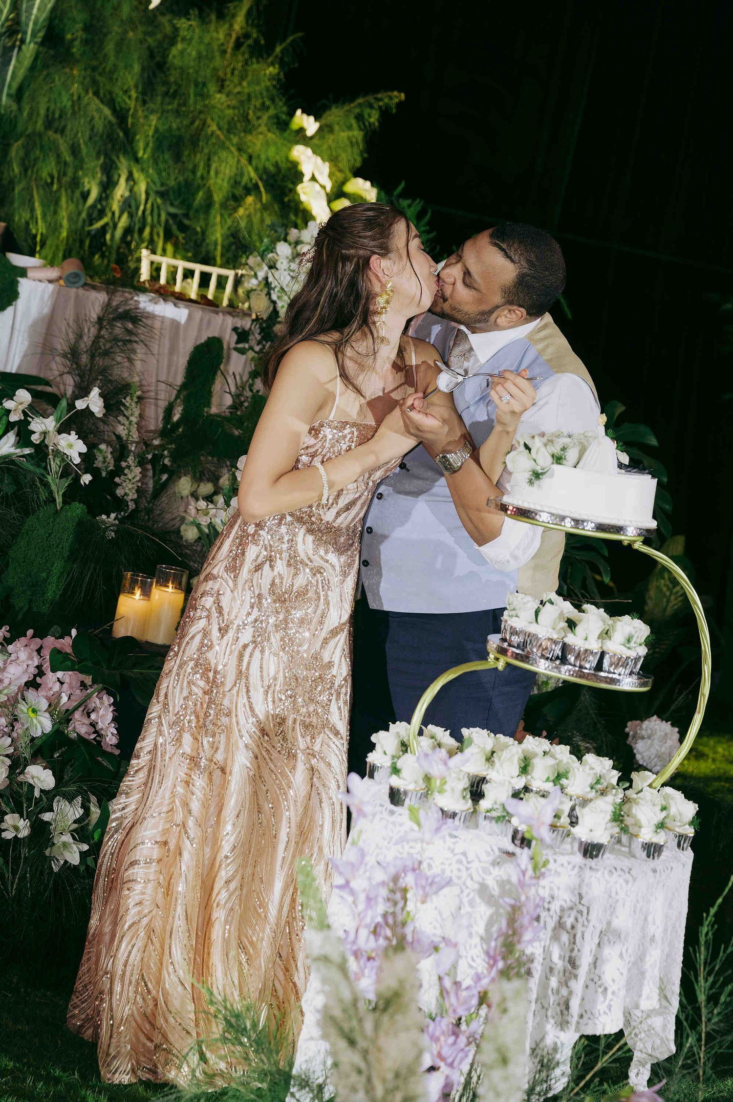 Newlyweds sharing a kiss beside tiered wedding cake and floral arrangements at a romantic outdoor garden reception.
