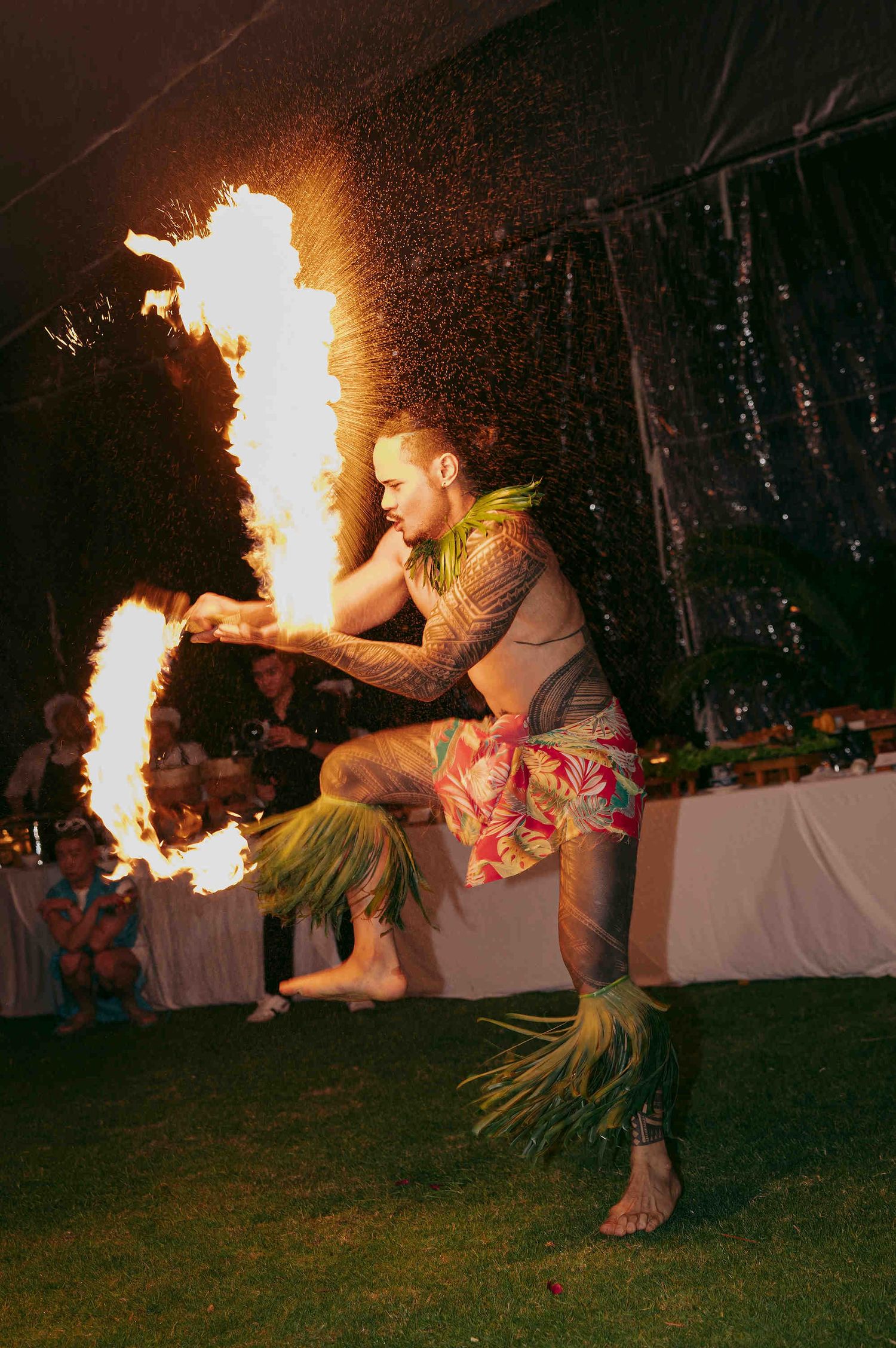 Tattooed fire dancer performs at outdoor luau event, breathing flames while wearing traditional grass skirt costume at night.