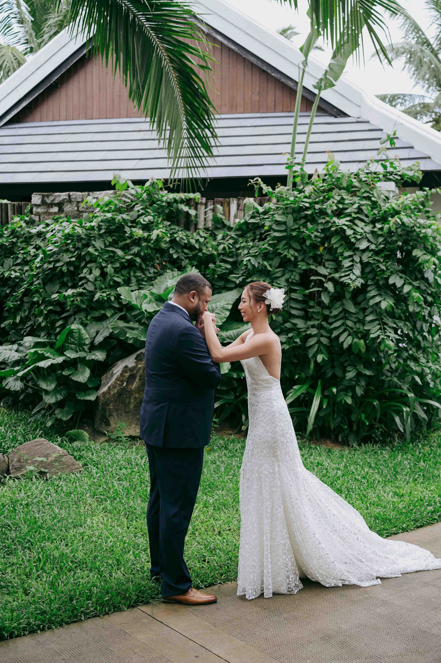 Bride and groom share a tender first look moment in a lush tropical garden, with palm trees and a wooden building backdrop.