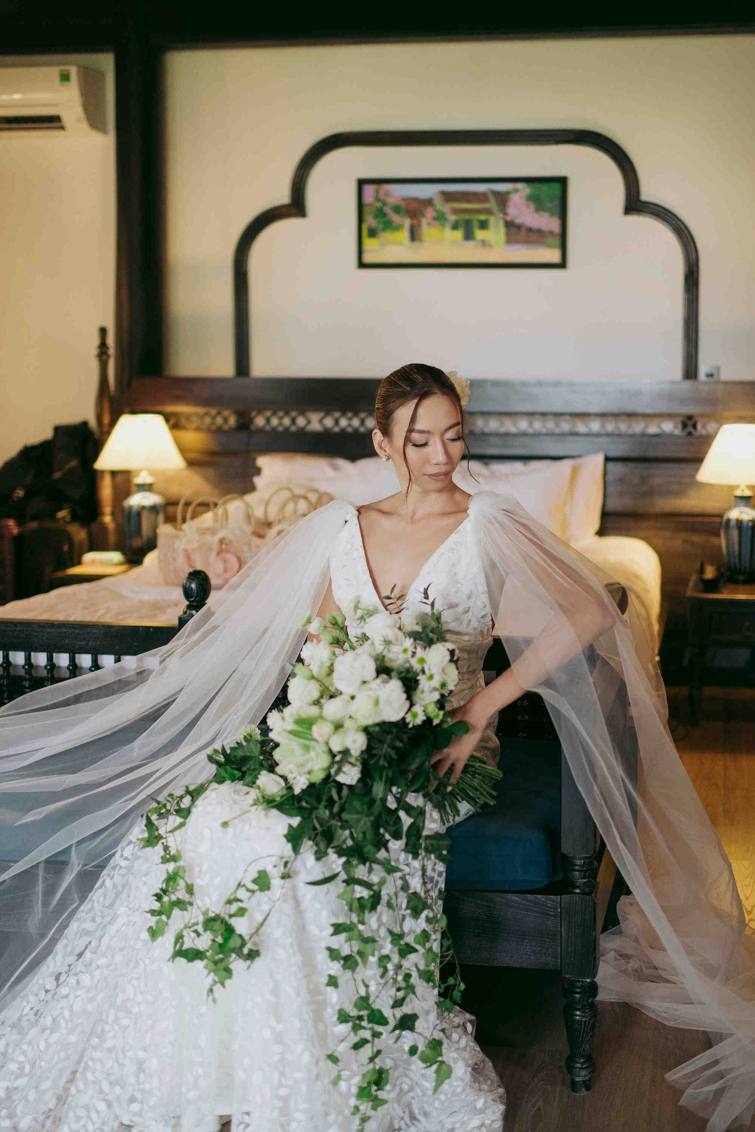 Bride in white lace gown with flowing veil holds cascading white bouquet while seated in elegant hotel room.