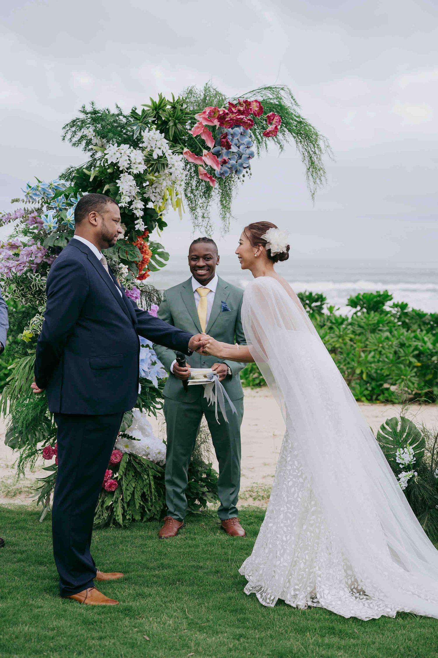 Couple exchanging vows at a tropical beach wedding ceremony with a colorful floral arch and officiant.