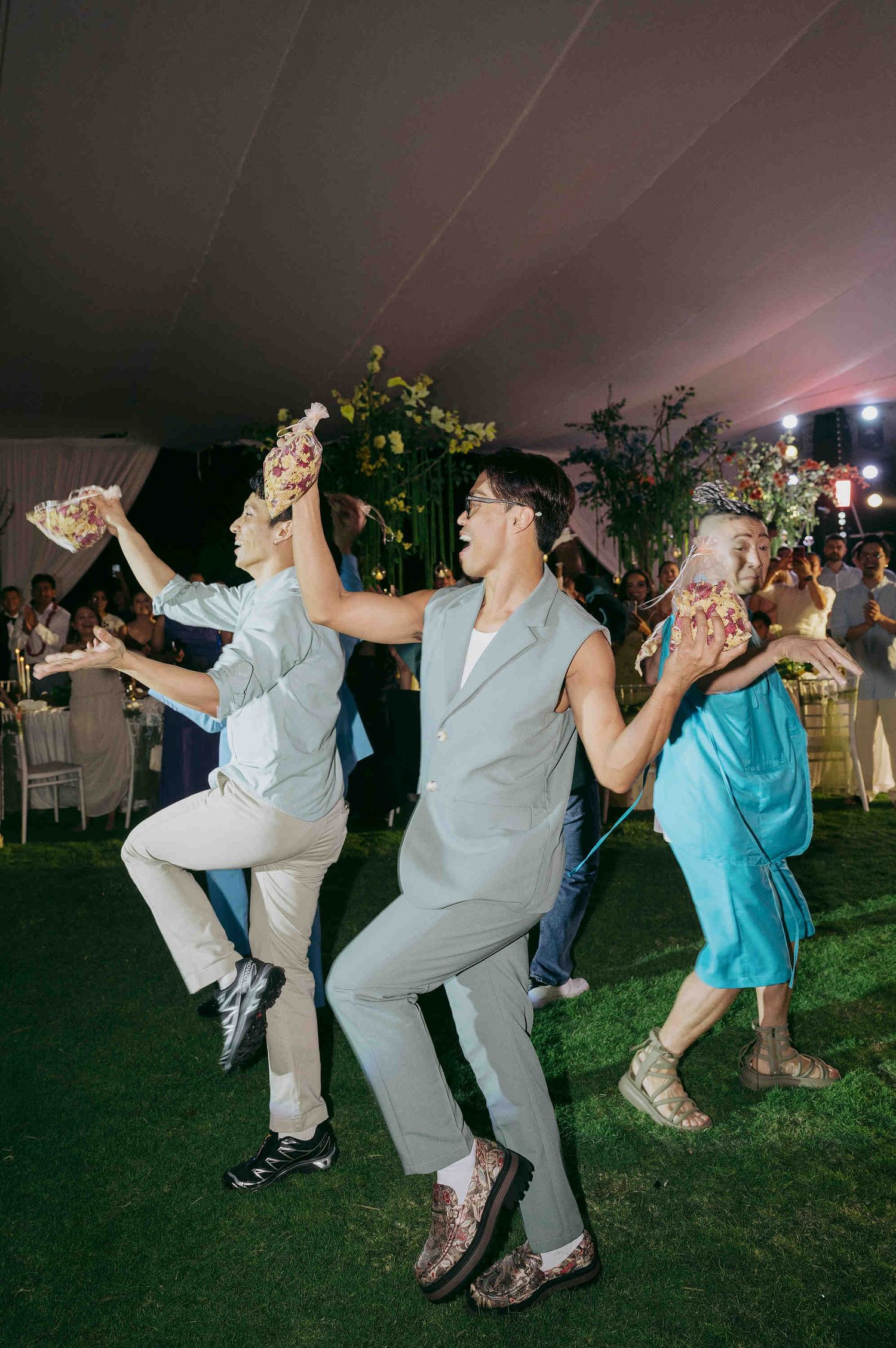 Guests dancing joyfully at an outdoor wedding reception under a tent with floral decorations and green landscaping.