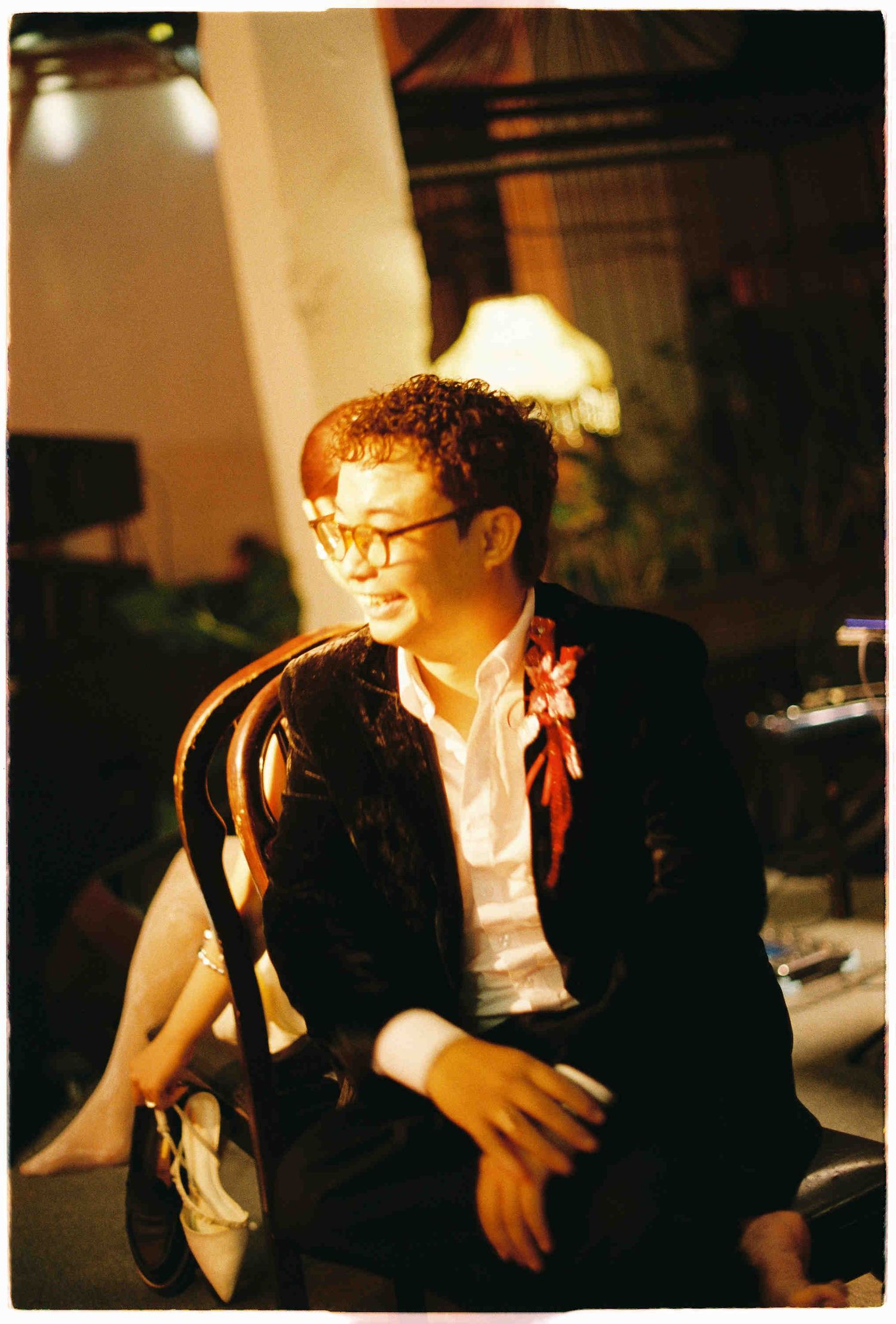 A young man in glasses and dark blazer with floral accessory sits in a wooden chair at an indoor evening event.