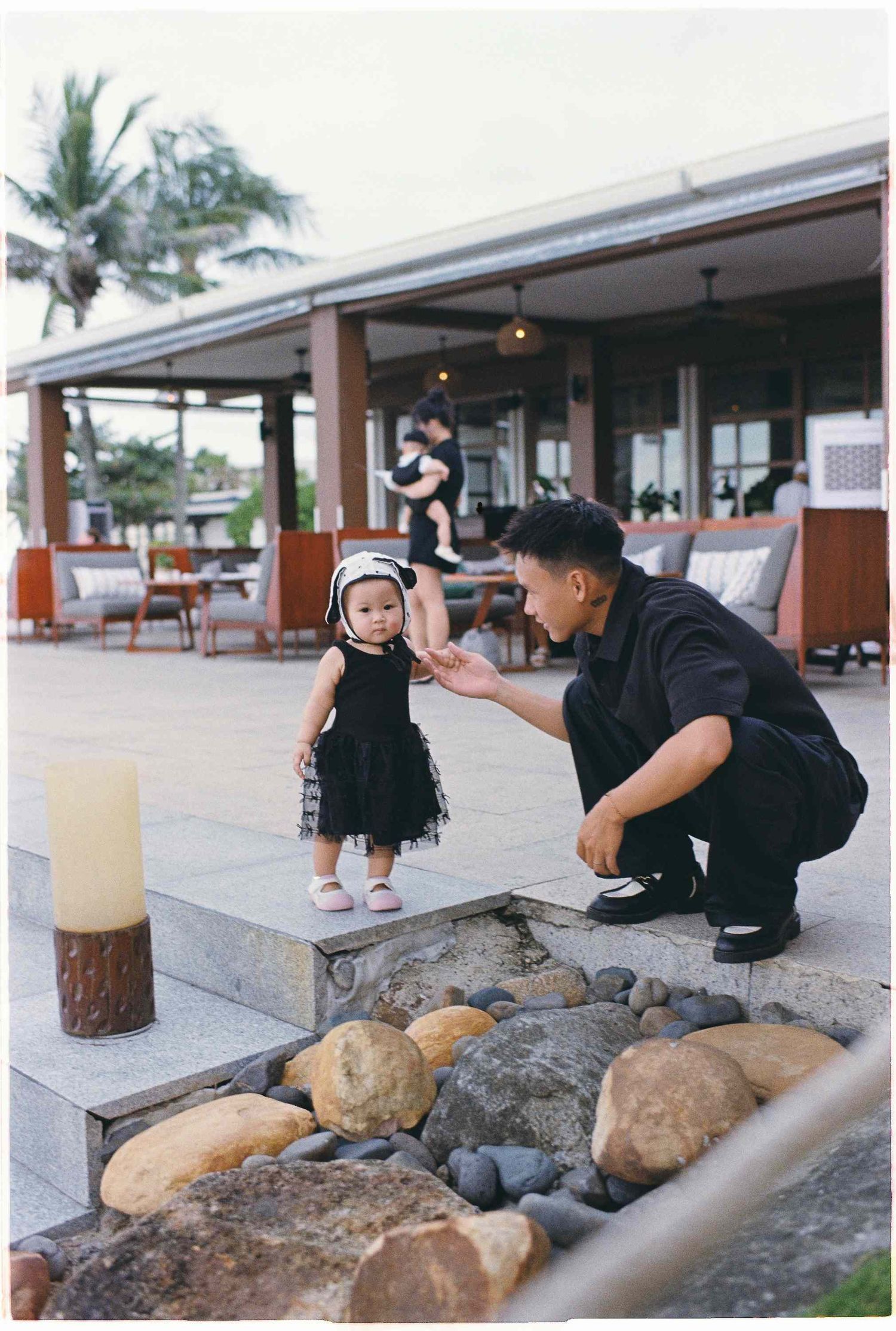 Da Nang family: toddler in a black dress stands on stone steps while an adult crouches nearby at an outdoor restaurant, by AnhVuWedding.