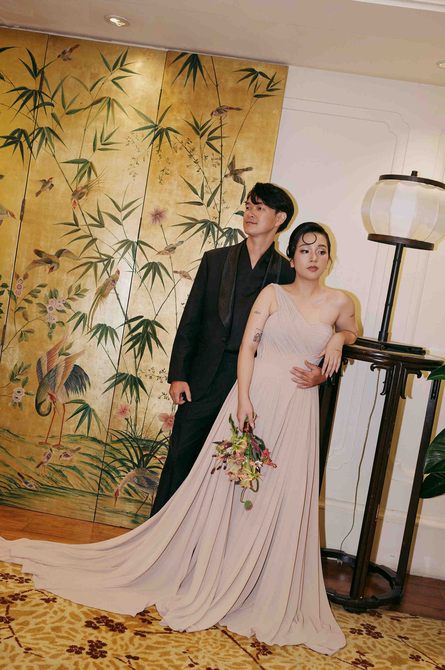 Couple posing in elegant attire before golden bamboo mural, bride in blush gown with bouquet, groom in black suit.