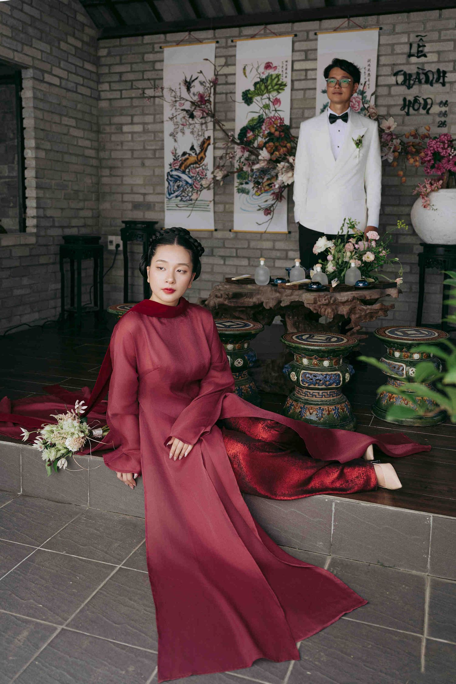 Vietnamese couple in traditional wedding attire, bride in red ao dai seated, groom in white suit standing in elegant venue.