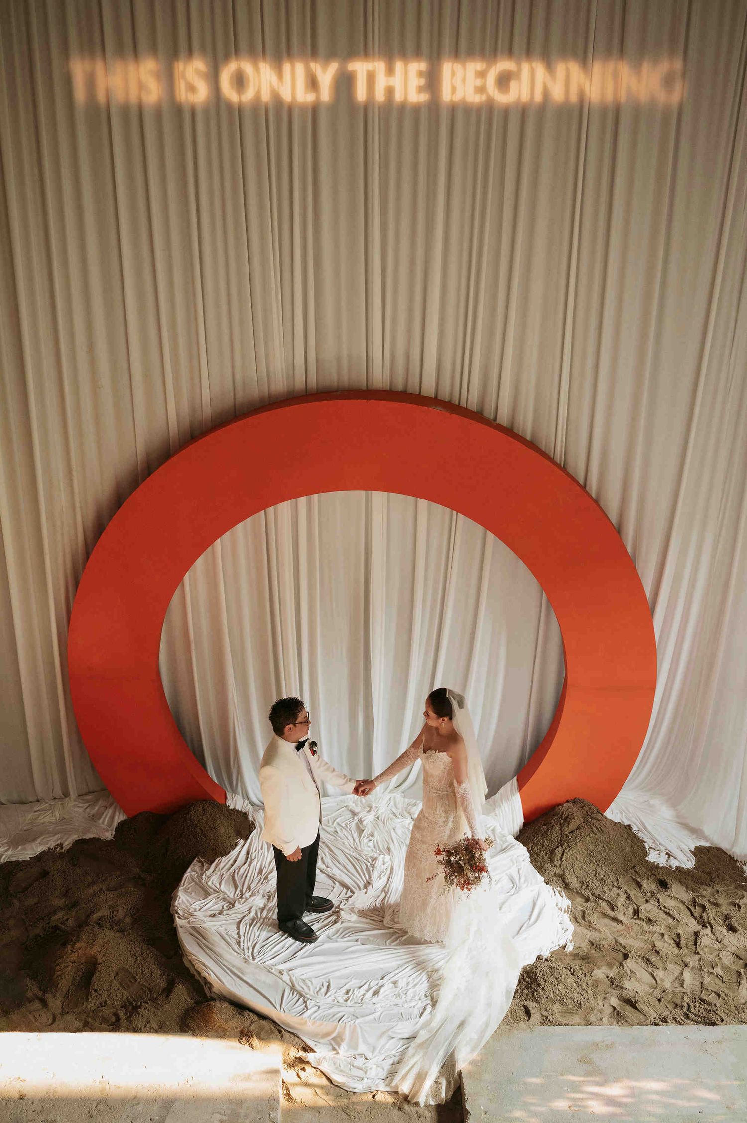 Bride and groom holding hands under a red arch with neon sign reading This Is Only The Beginning.