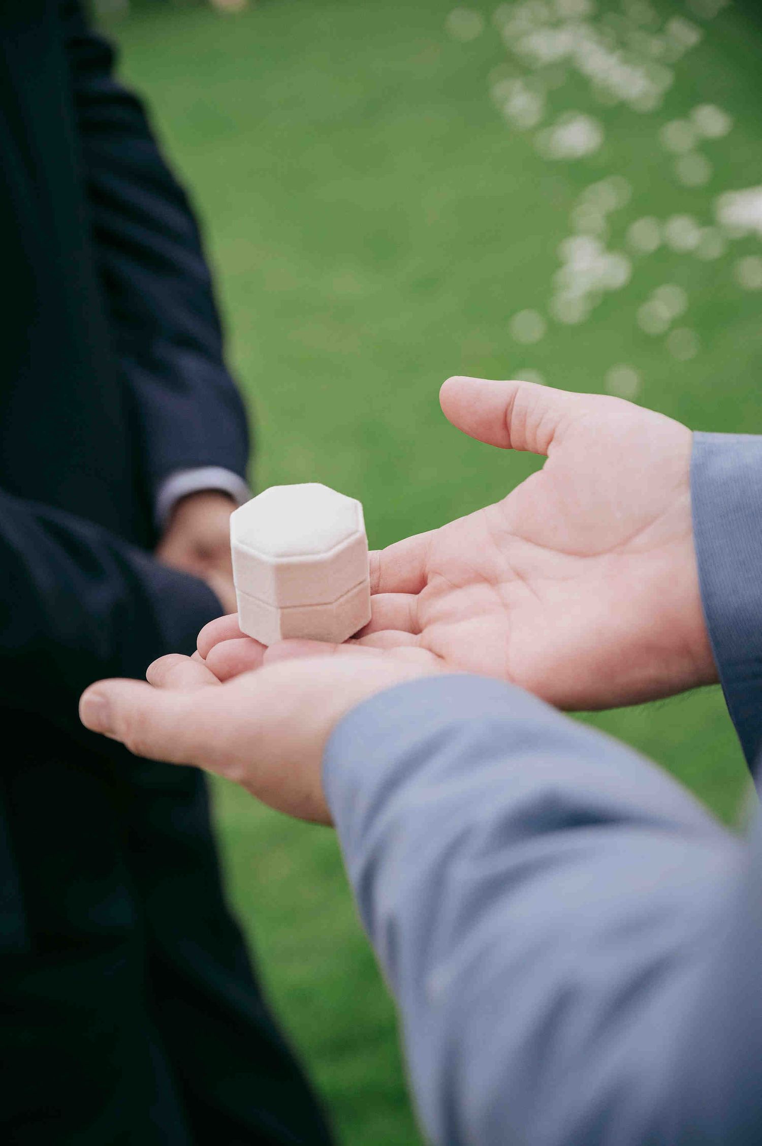Groom holding a small octagonal white ring box during an outdoor wedding ceremony on green grass.