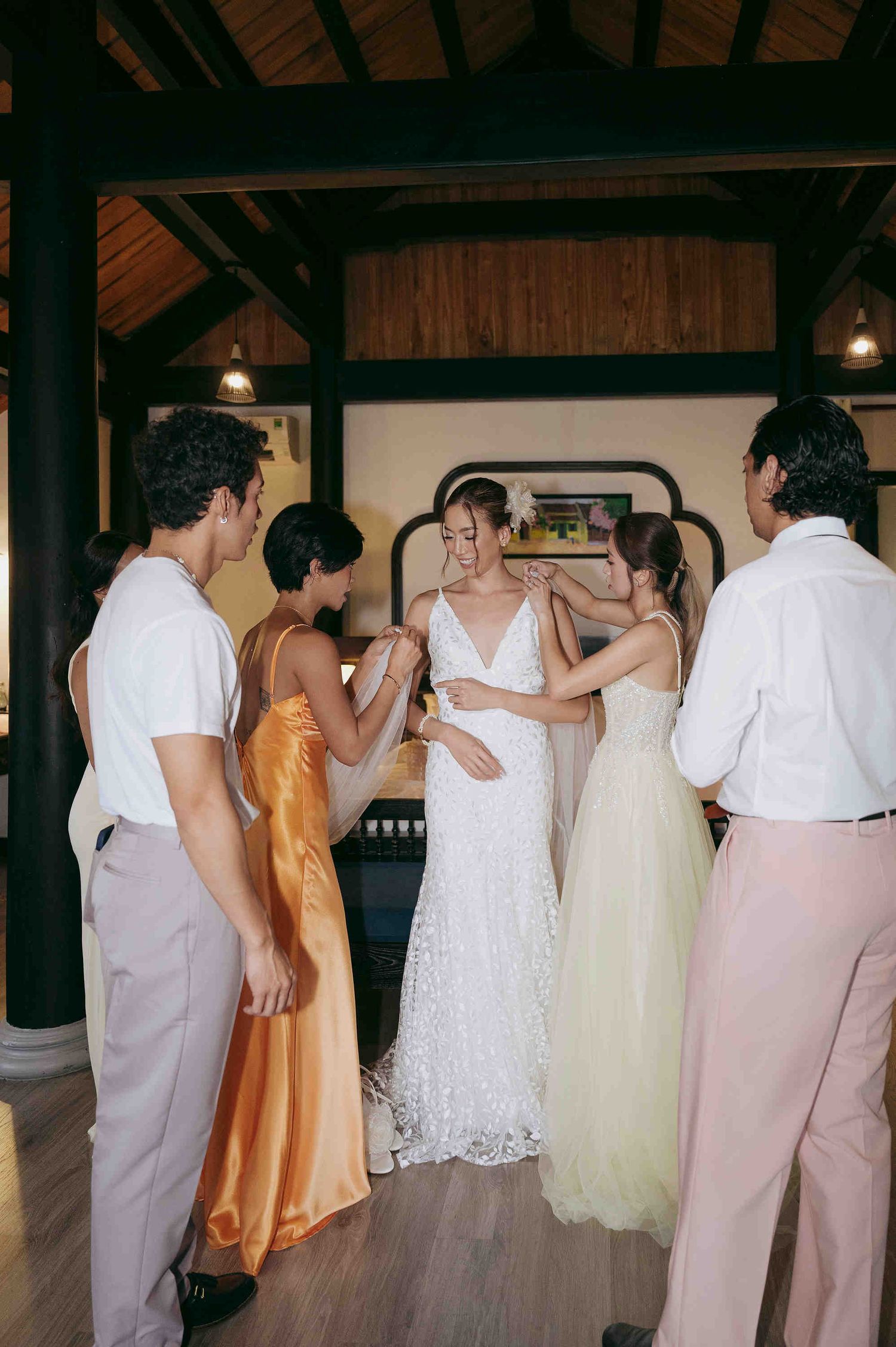 Bride in white lace gown surrounded by bridesmaids and groomsmen helping her prepare in elegant venue.