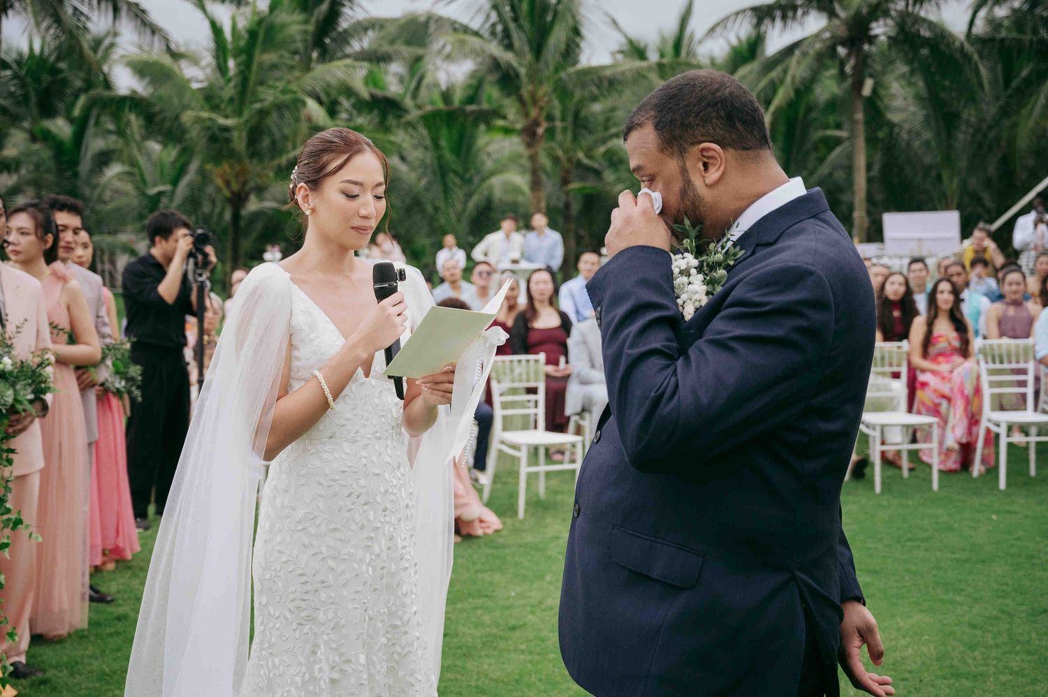 Bride reads wedding vows to emotional groom at outdoor ceremony, guests seated in white chairs surrounded by tropical greenery.