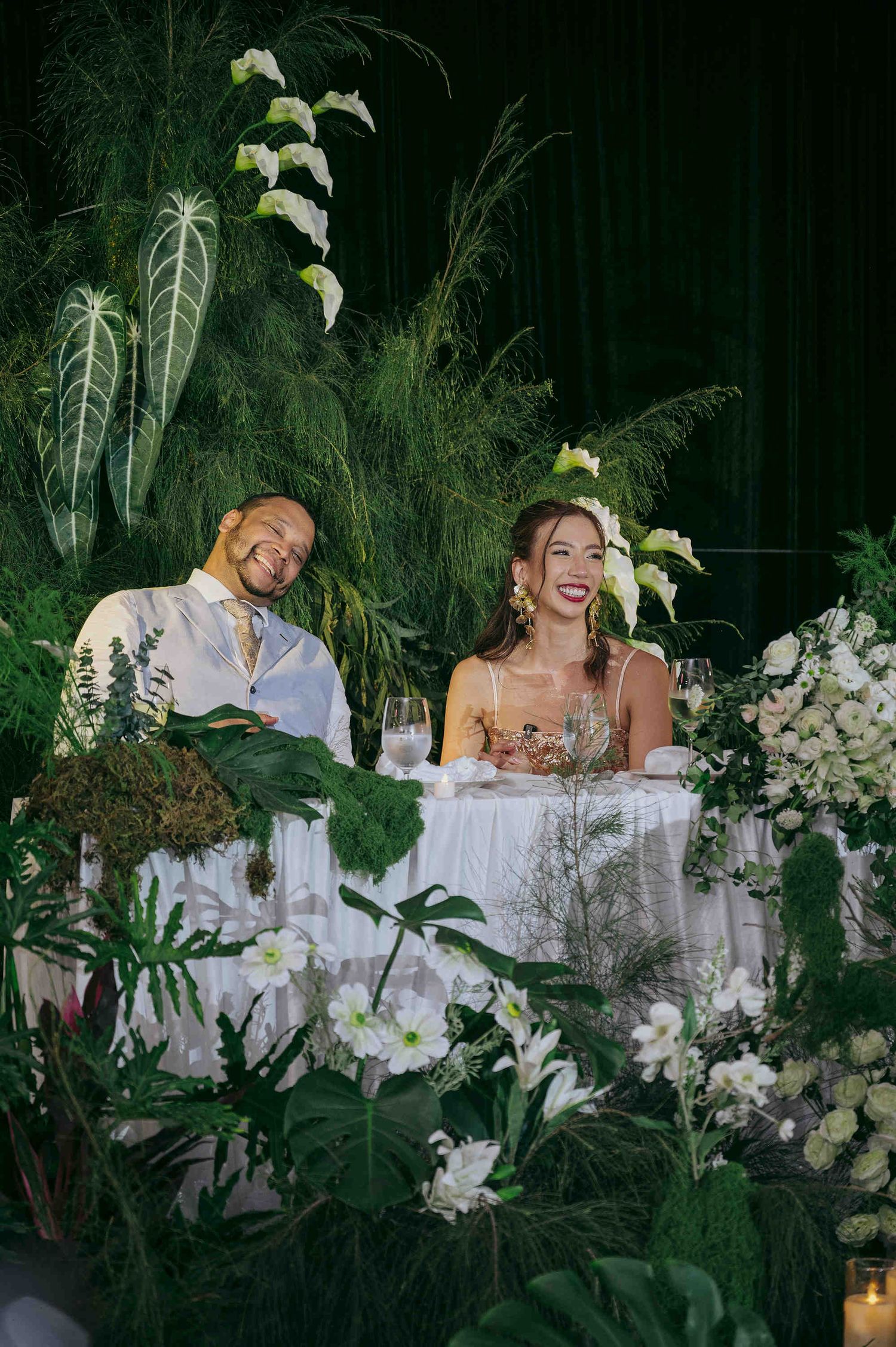 Couple seated at floral sweetheart table surrounded by lush tropical greenery and white flowers at wedding reception.