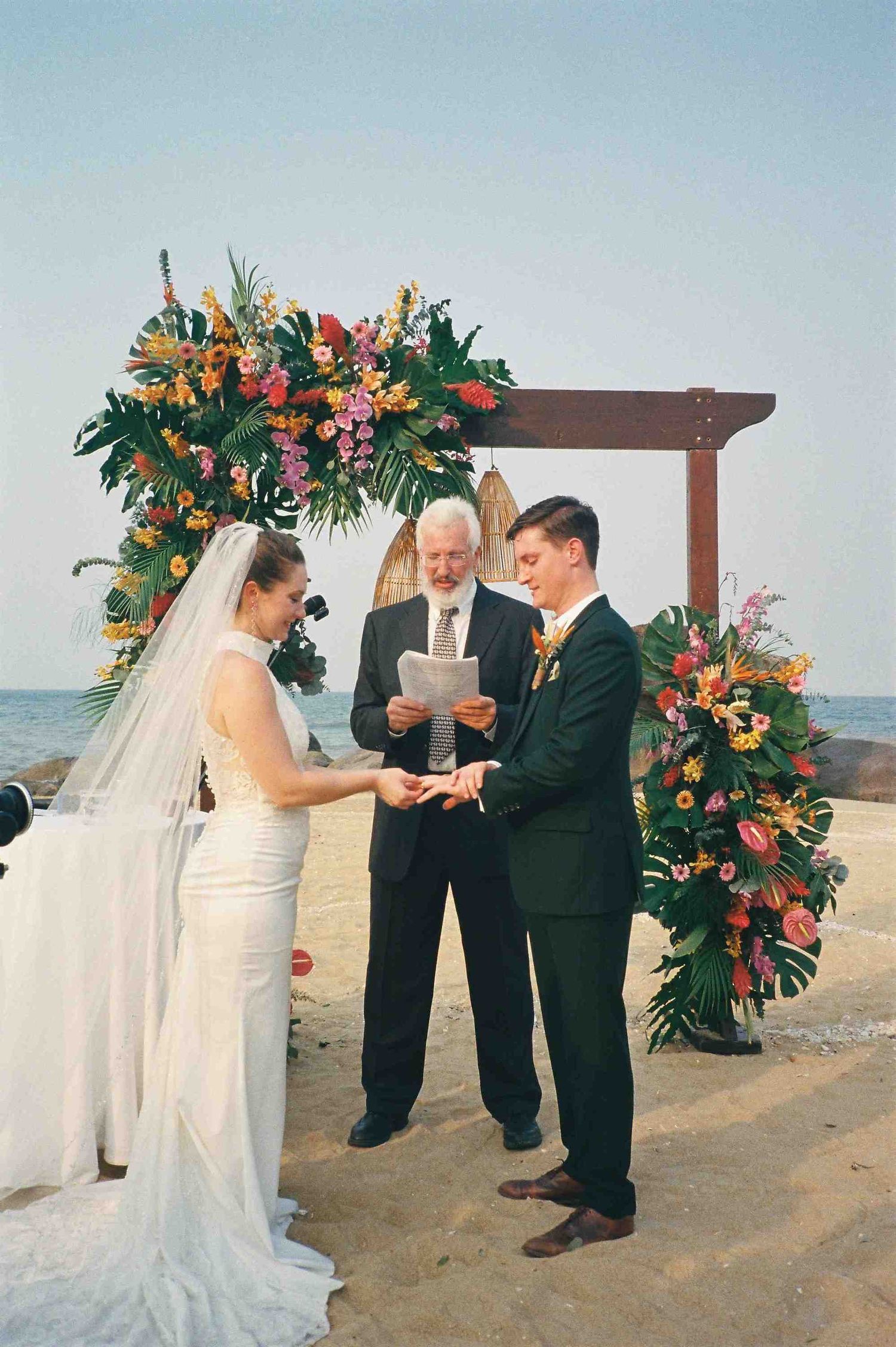 Lang Co wedding ceremony: beach wedding ceremony under a wooden arch decorated with vibrant tropical flowers, by AnhVuWedding.
