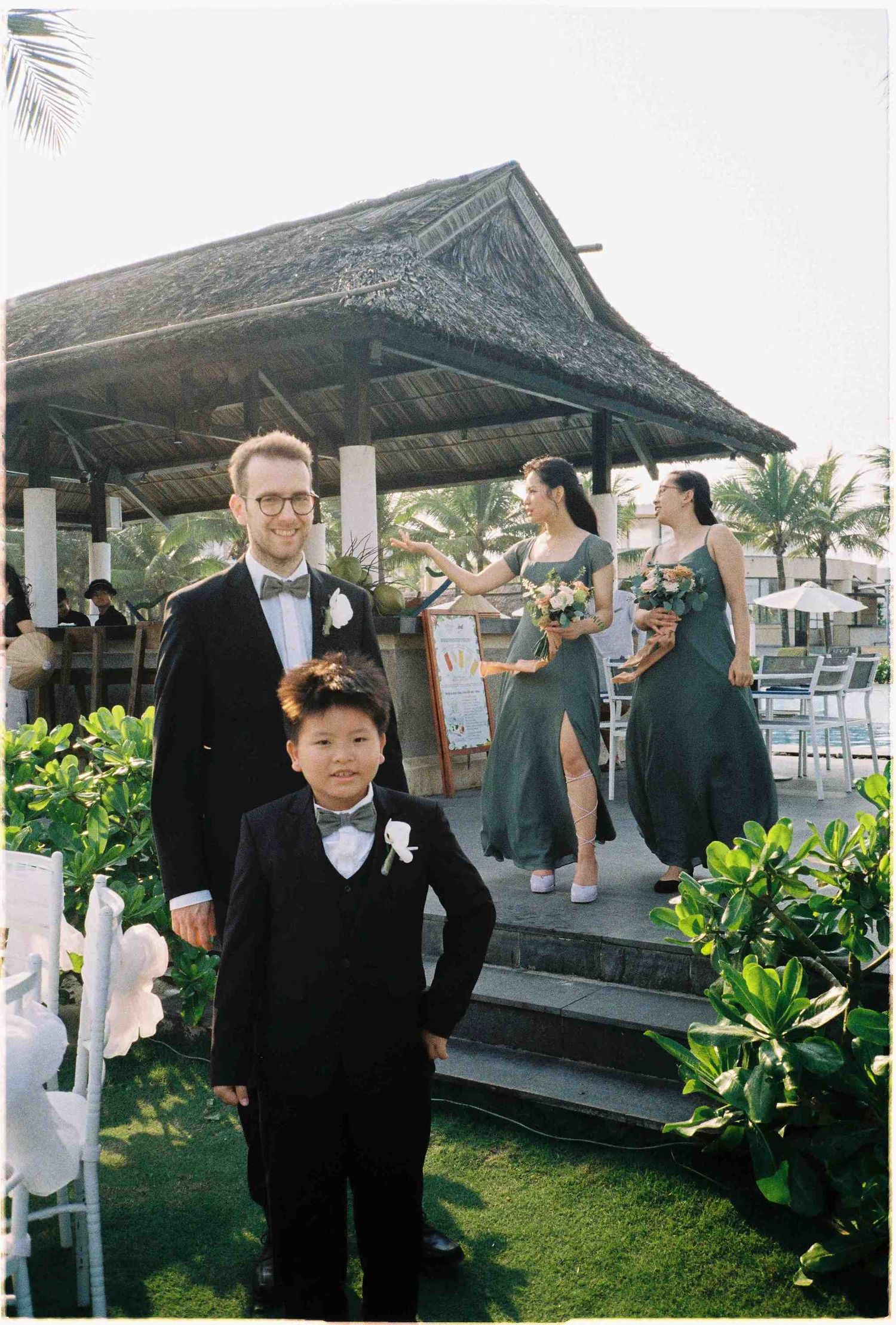 A groomsmen and ring bearer in black tuxedos pose outdoors at a tropical wedding with bridesmaids in green gowns.