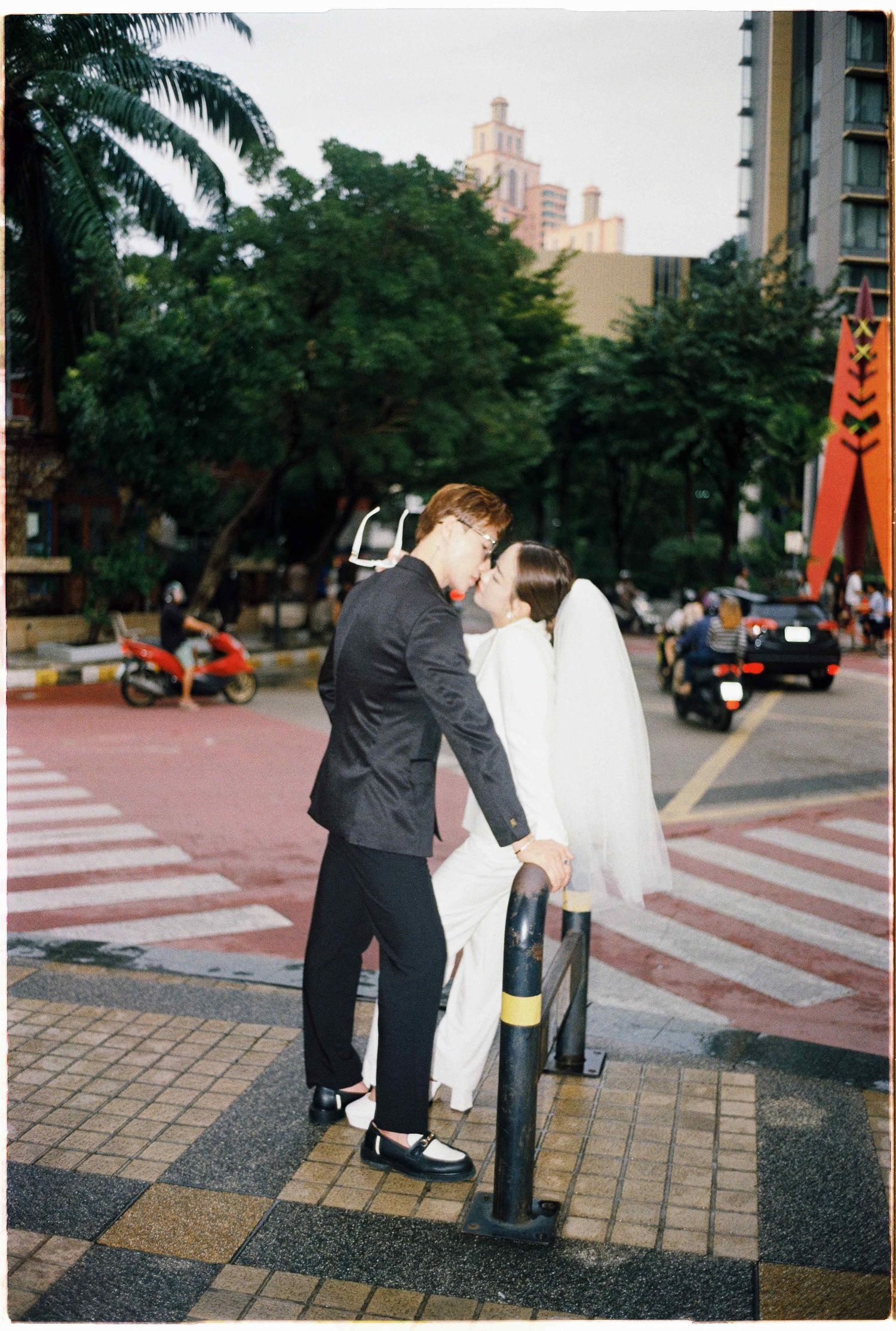 Saigon pre-wedding: couple in black and white attire interact on a paved urban plaza with palm trees in background, by AnhVuWedding.