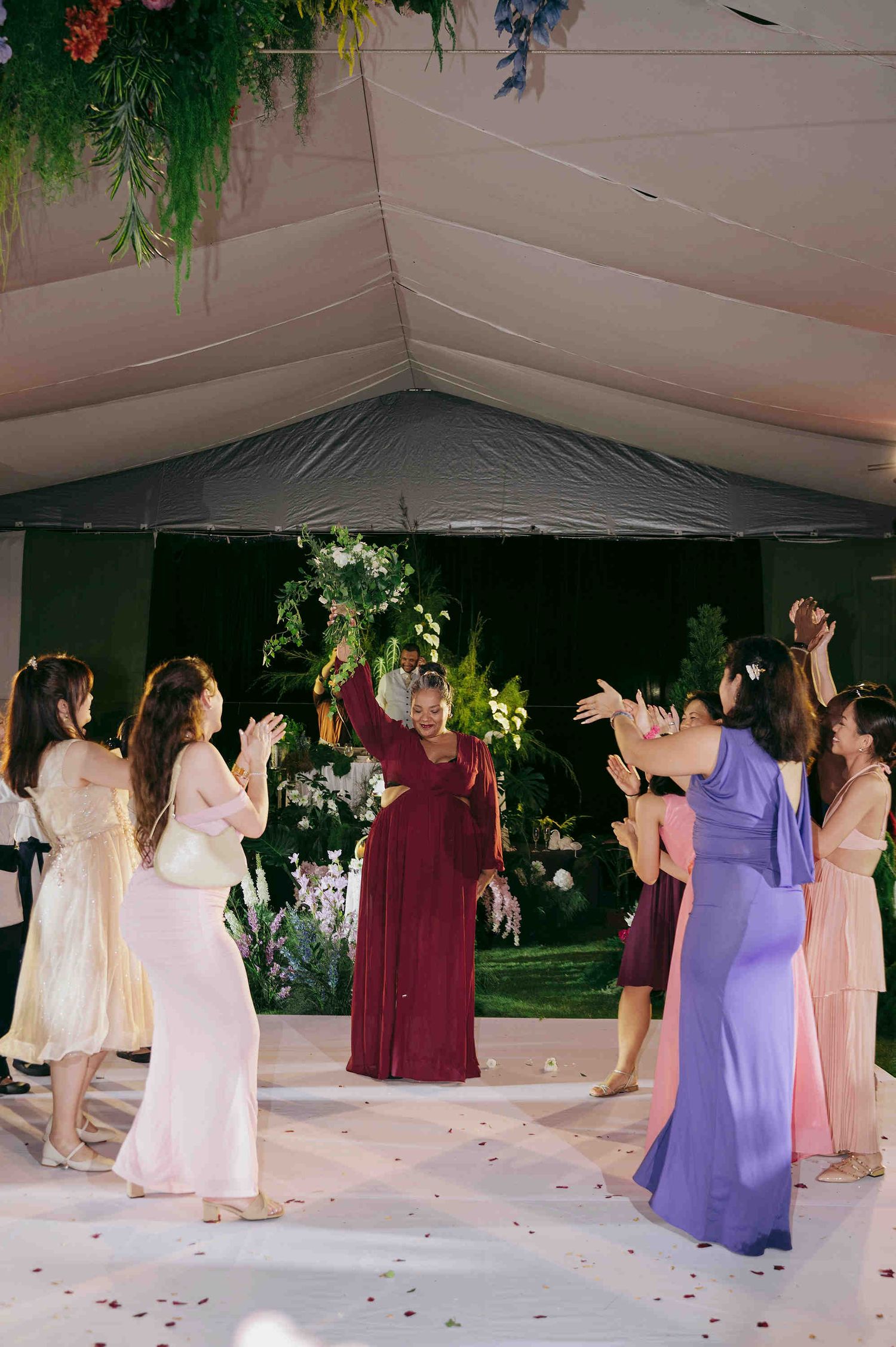 Women in colorful formal dresses celebrating on a dance floor at an outdoor wedding reception under a tent.