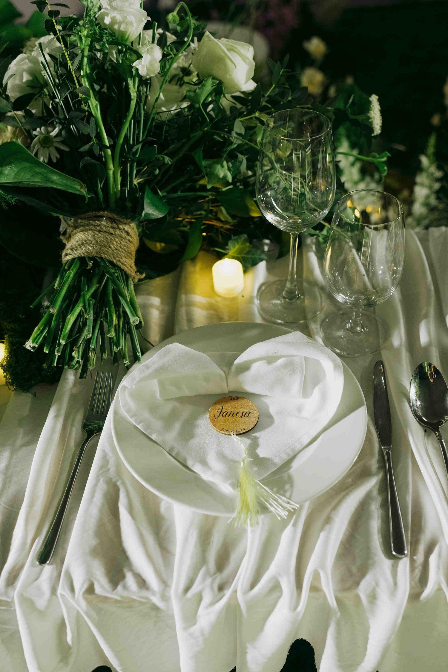 Elegant wedding place setting with white napkin, gold name card, crystal glasses, and floral arrangement.