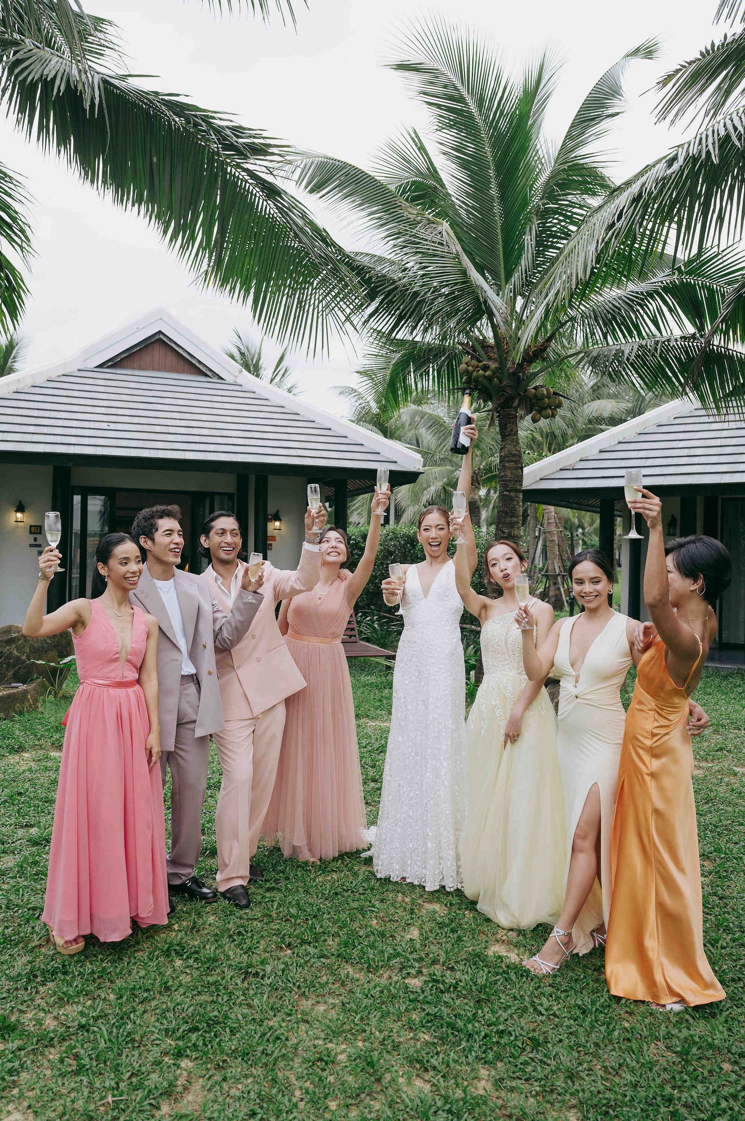 Wedding party toasting with champagne outdoors, bridesmaids in colorful gowns and bride in white, tropical palm trees background.