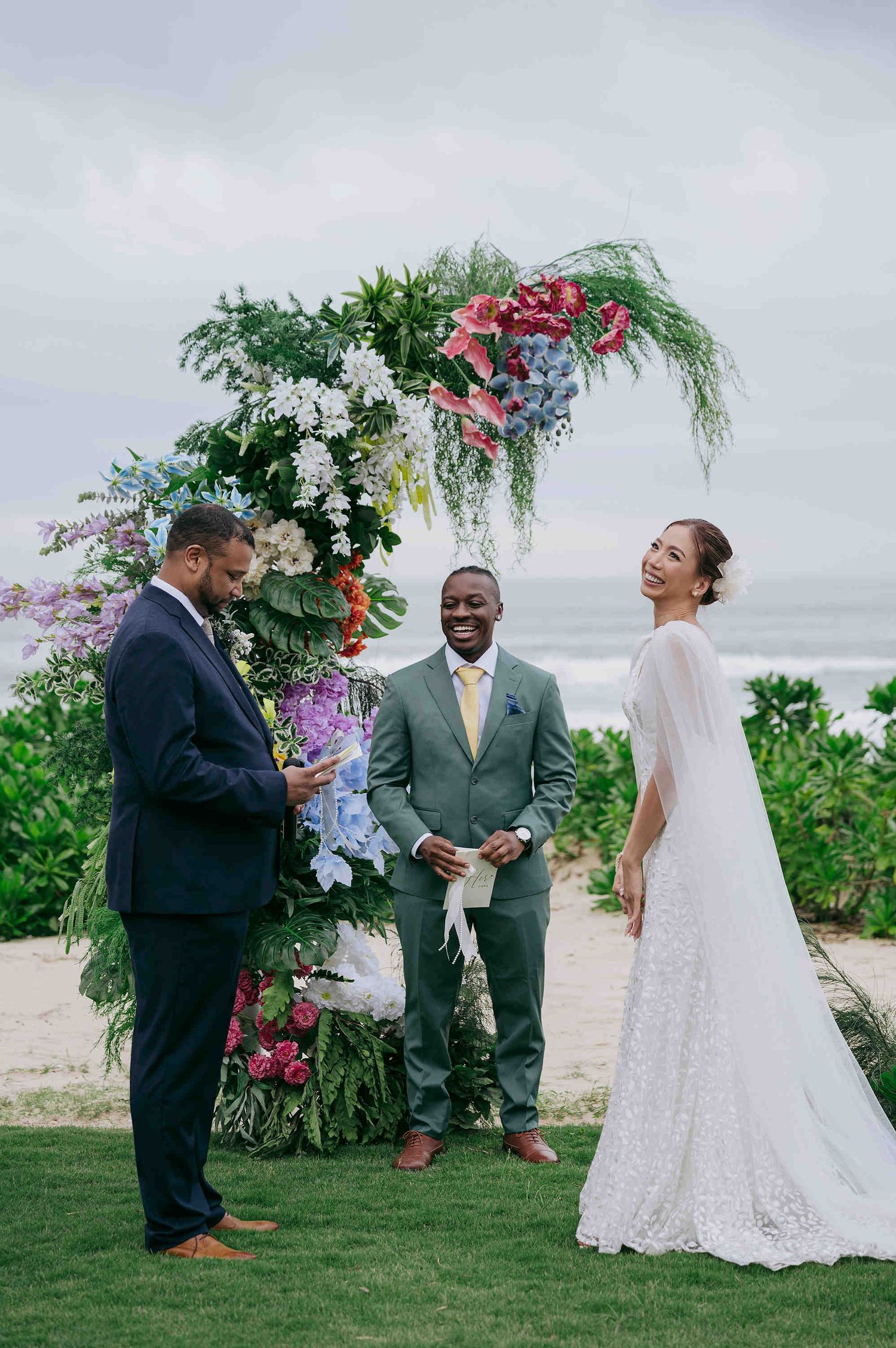 Couple exchanging vows at a tropical beach wedding ceremony with colorful floral arch and officiant.