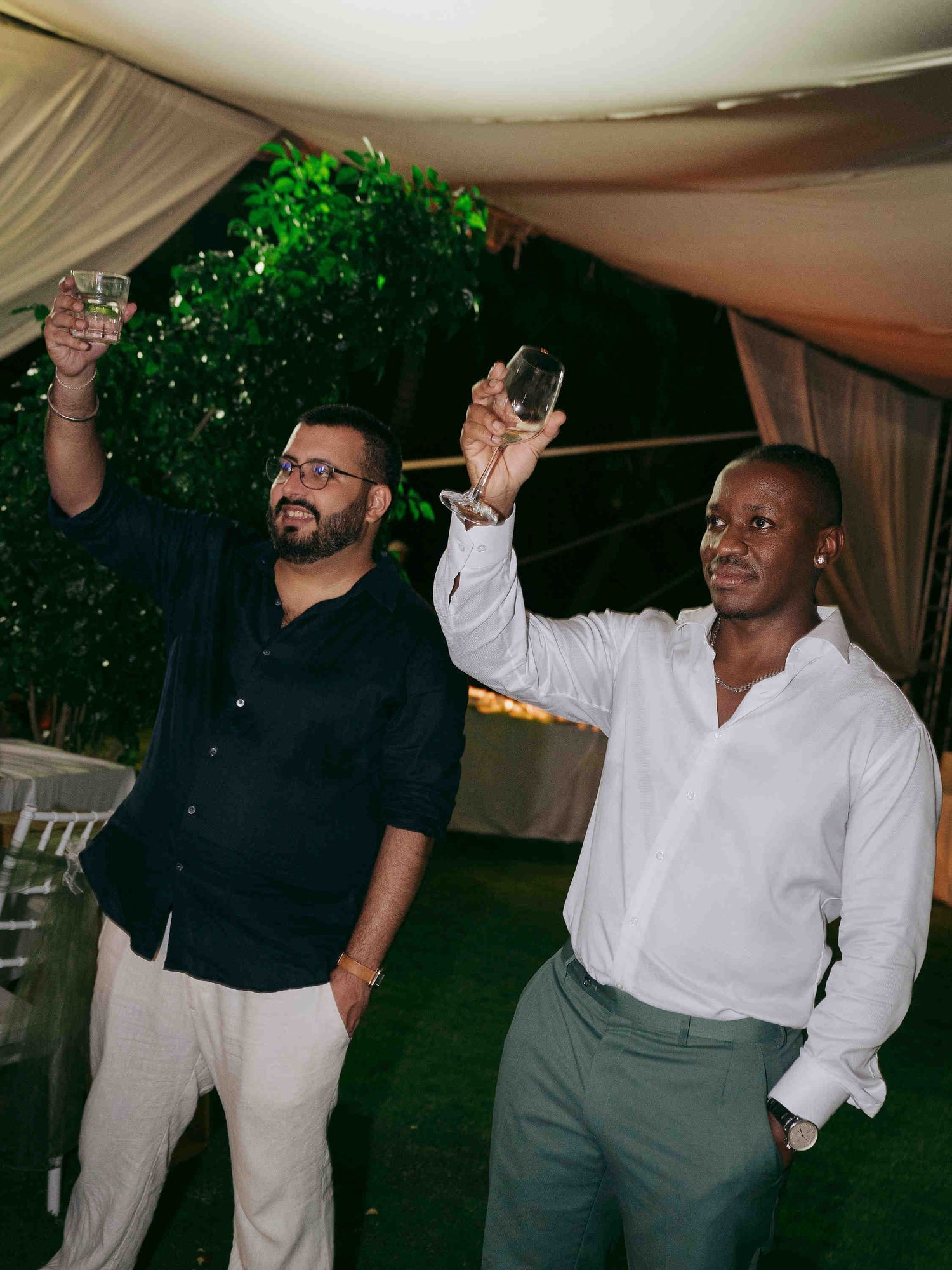 Two men raising glasses in a toast at an outdoor evening event under a white tent.