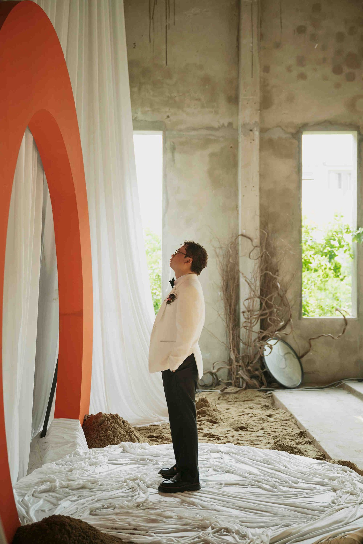 Man in white sweater stands in rustic room with white drapes, orange arch, and natural light from open windows.