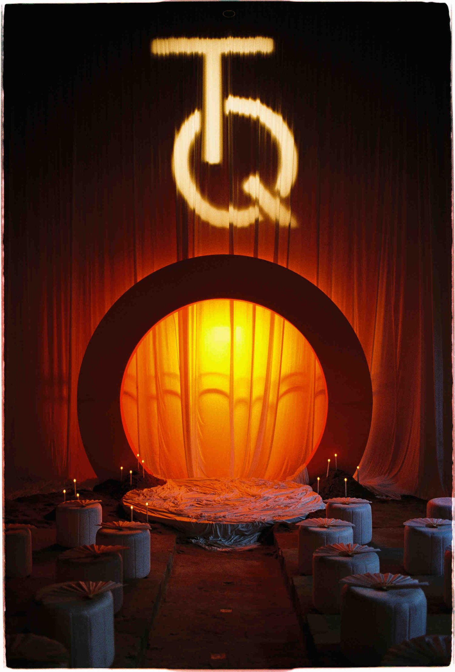 Art deco stage with glowing circular arch, TQ monogram in lights, candles, and white ottomans in dramatic orange ambiance.