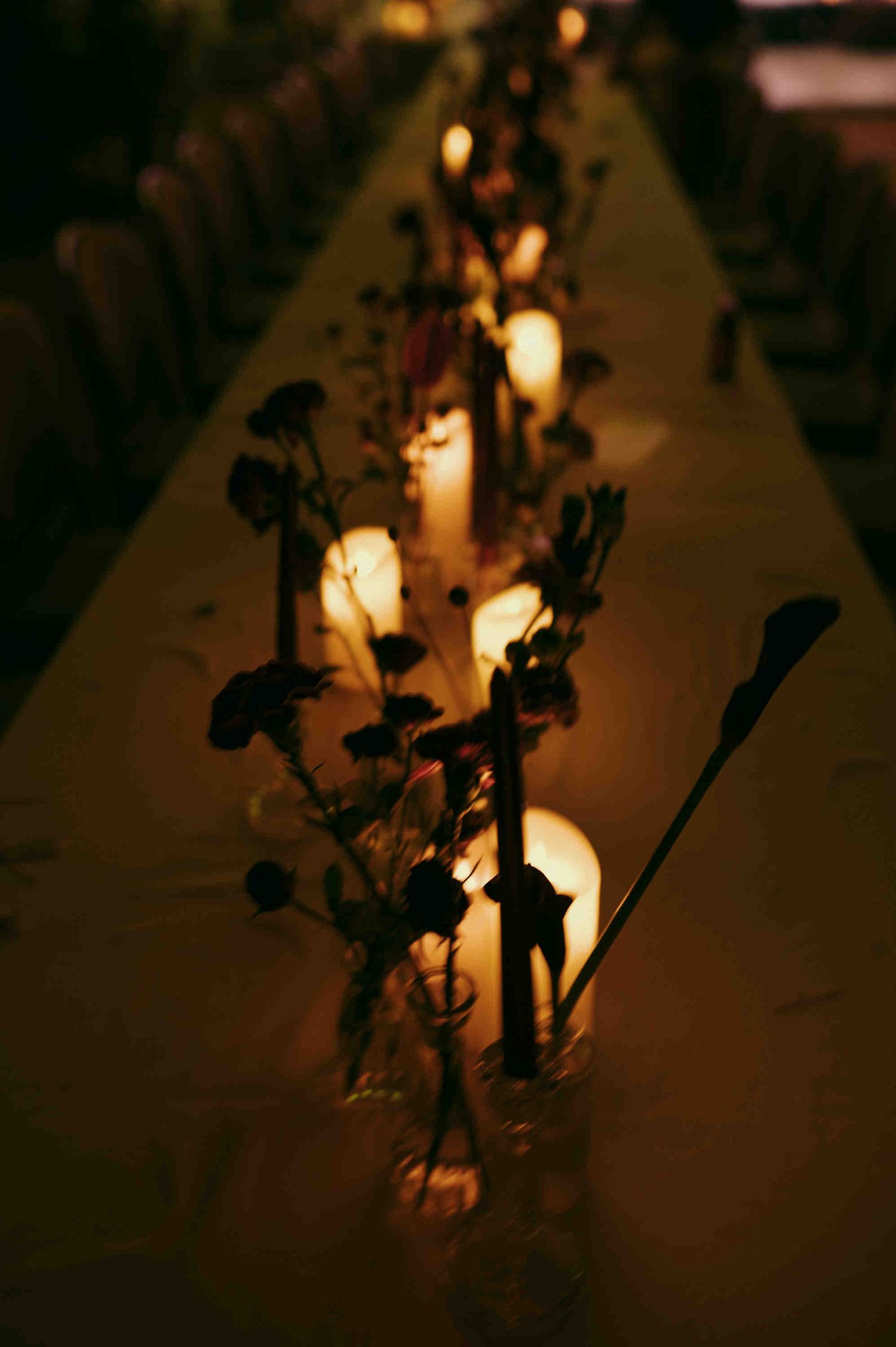 Romantic candlelit dinner table with glowing pillar candles and dark floral arrangements creating an intimate ambiance.