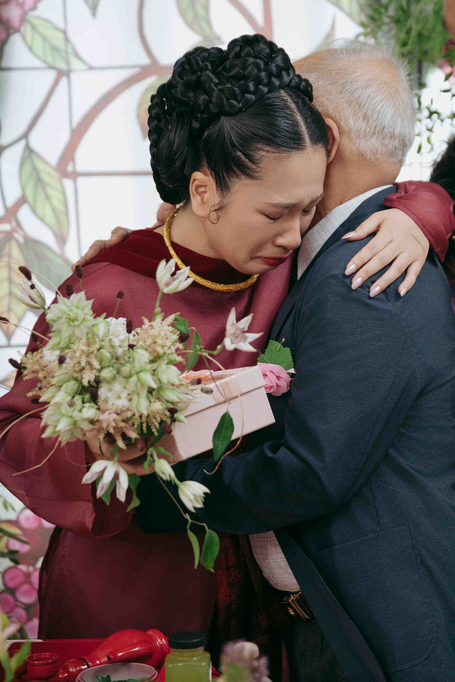 Emotional bride in red ao dai crying while embracing an elderly man, holding a bouquet at a wedding ceremony.