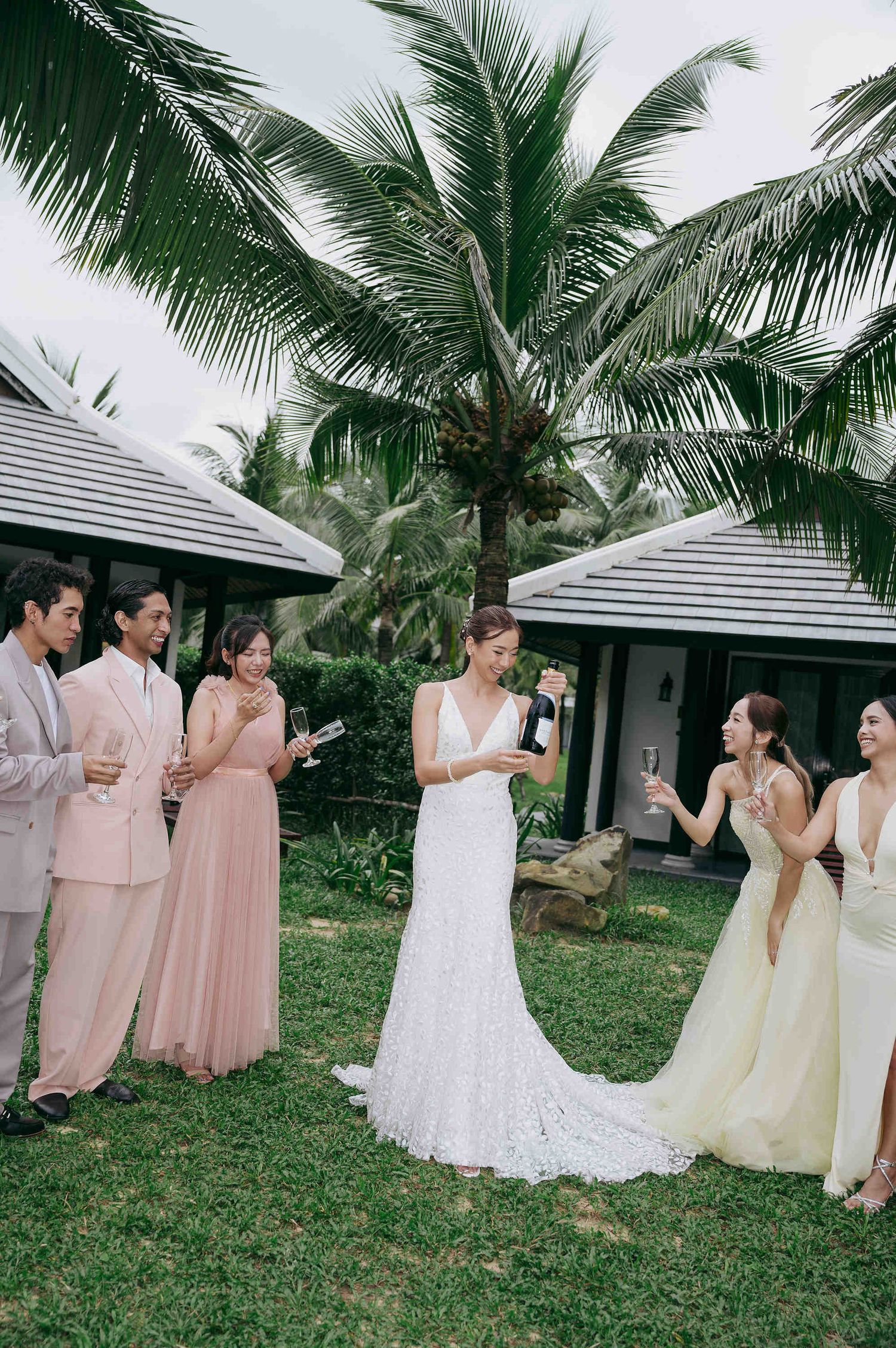 Bride in white lace gown opens champagne with bridesmaids in pink and yellow dresses outdoors near tropical palm trees.