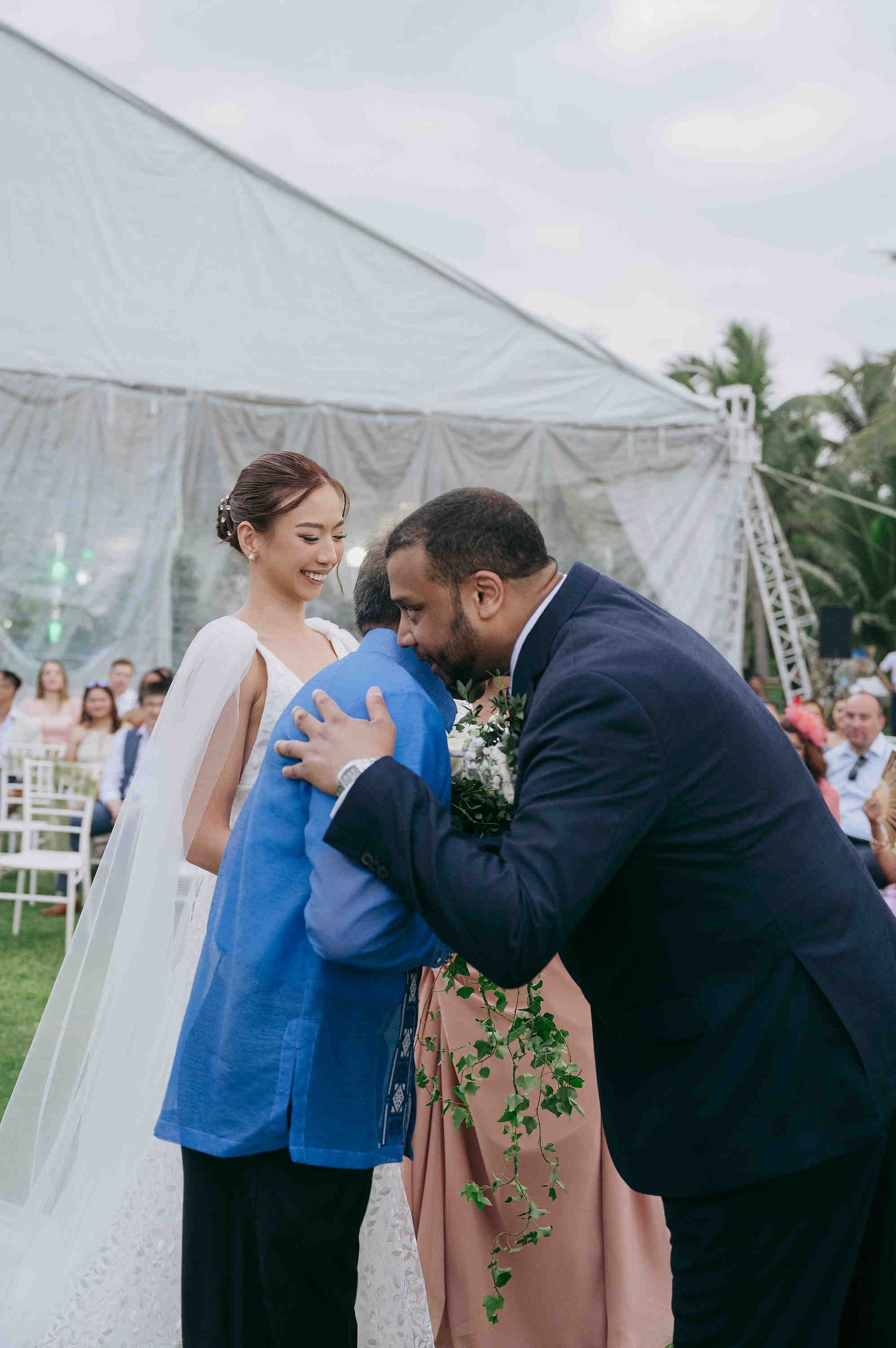 Groom in navy suit embracing family member at outdoor wedding ceremony while bride in white gown smiles nearby.
