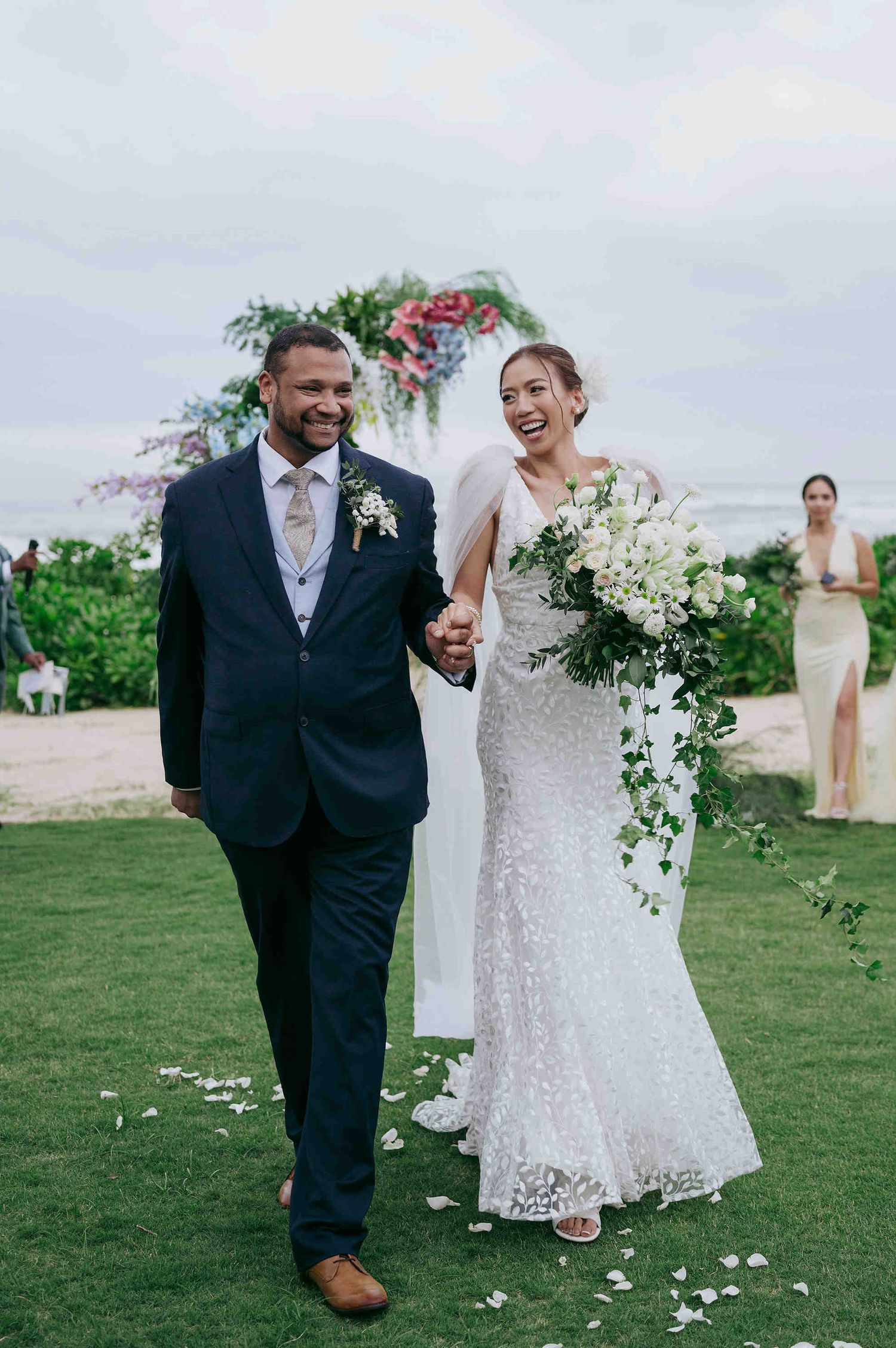 Smiling newlyweds walk down the aisle at an outdoor beach wedding, bride in lace gown holding white floral bouquet.