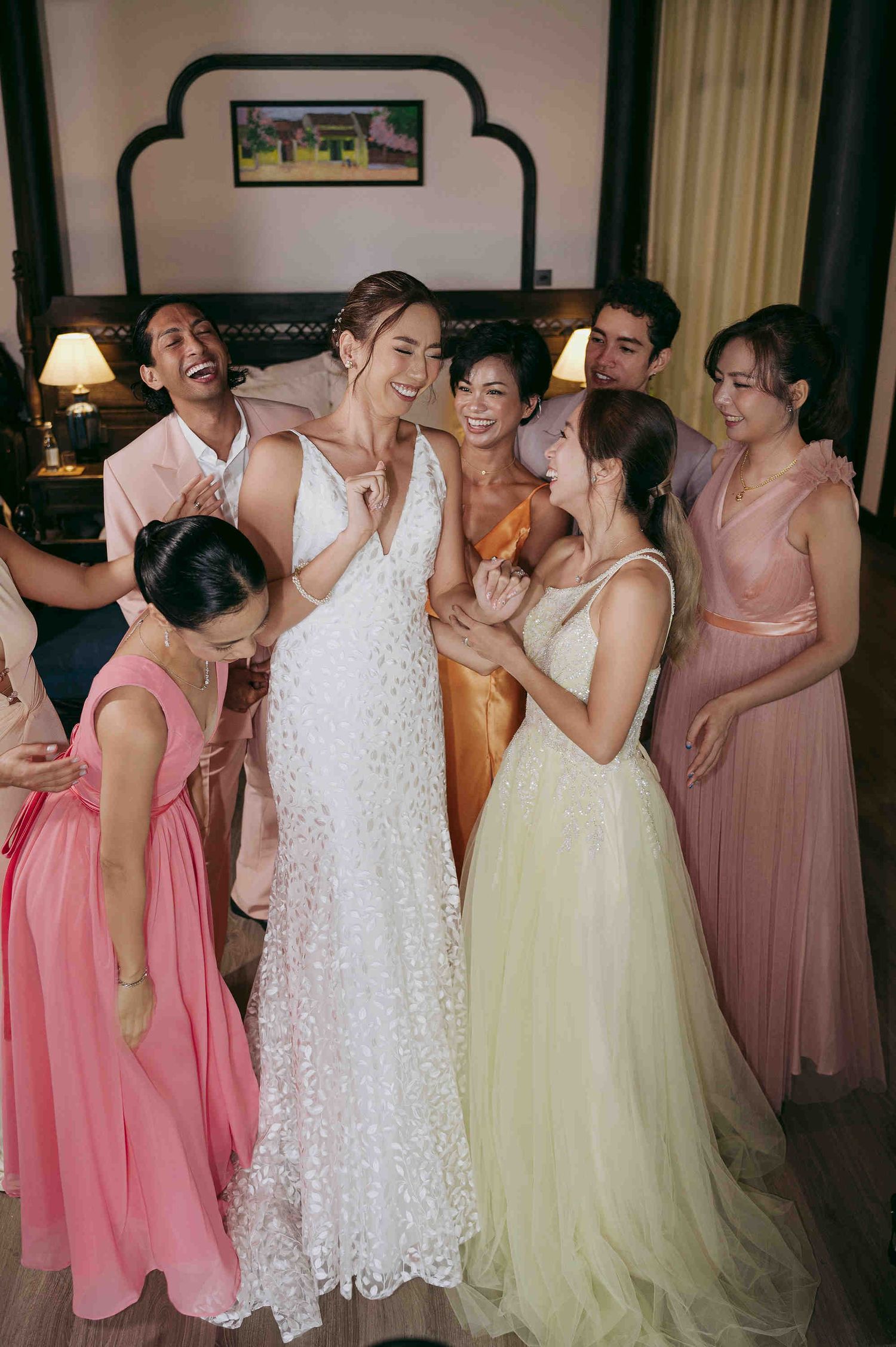 Bride in white lace gown surrounded by smiling bridesmaids in pastel dresses during wedding celebration indoors.