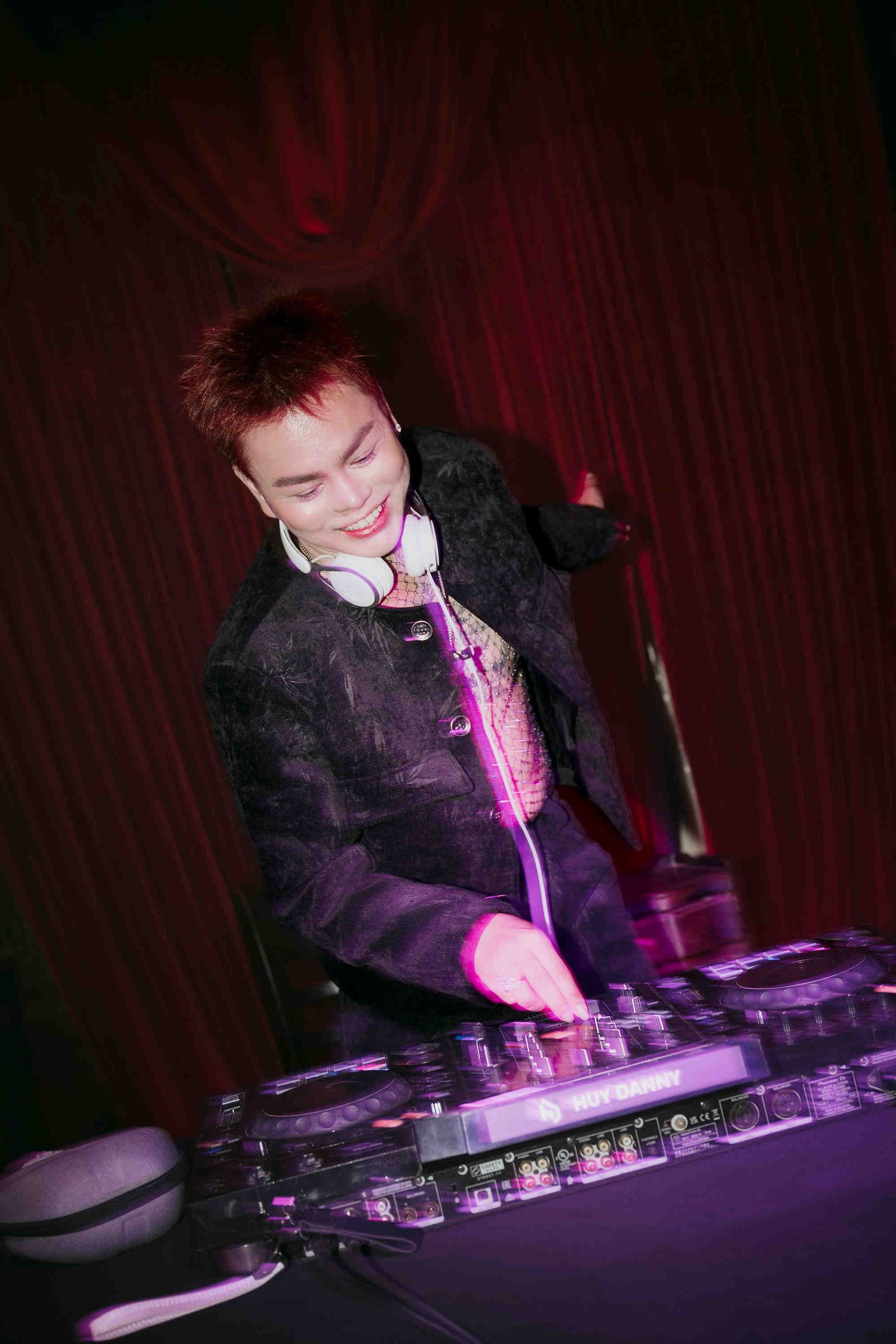 Smiling DJ with red hair and white headphones mixing tracks at a DJ booth under purple stage lighting.