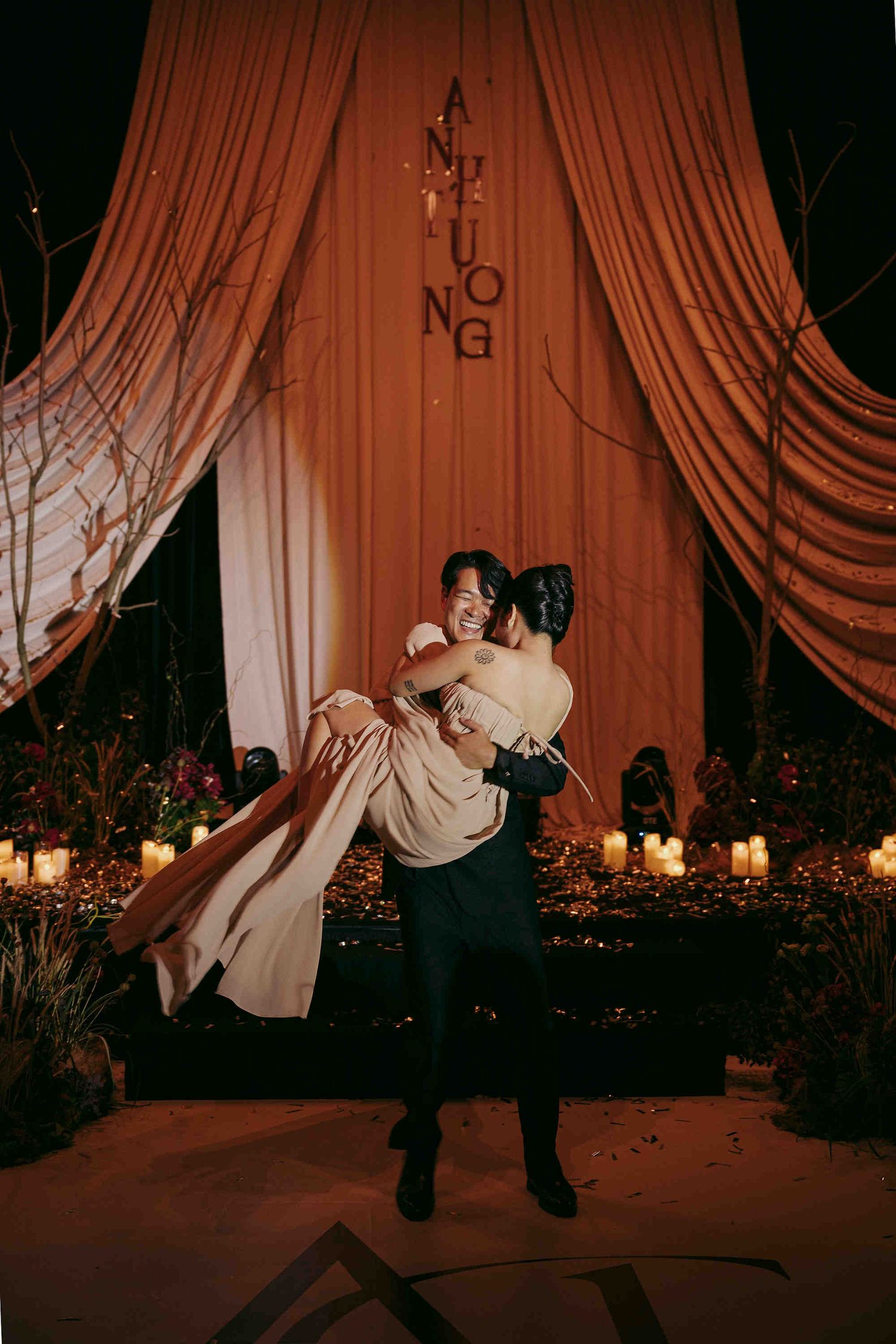 Groom lifting bride in elegant wedding reception with candlelit backdrop and orange draped curtains.