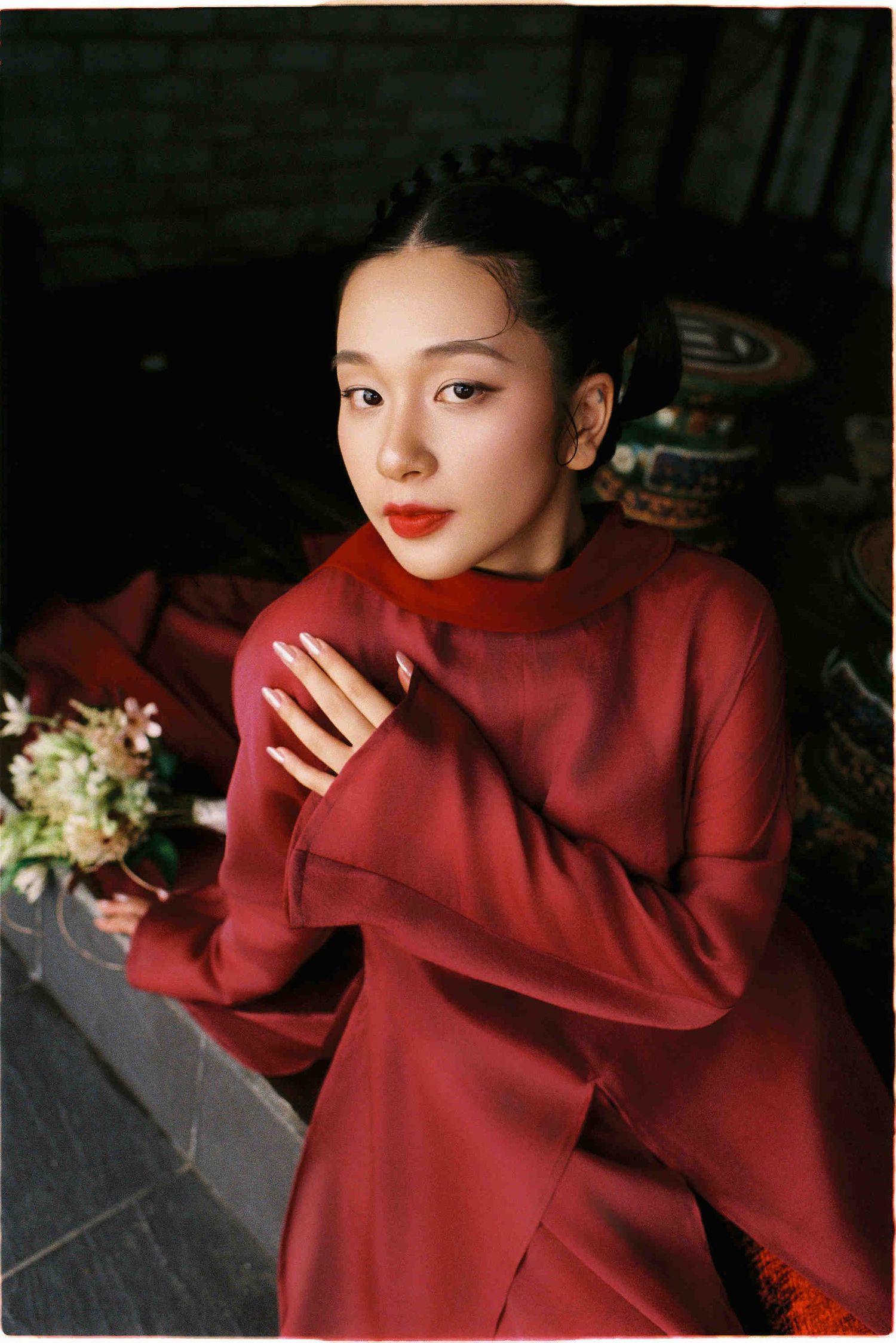 Young woman in elegant red dress with braided updo and red lips, posing gracefully in a traditional Asian-inspired setting.