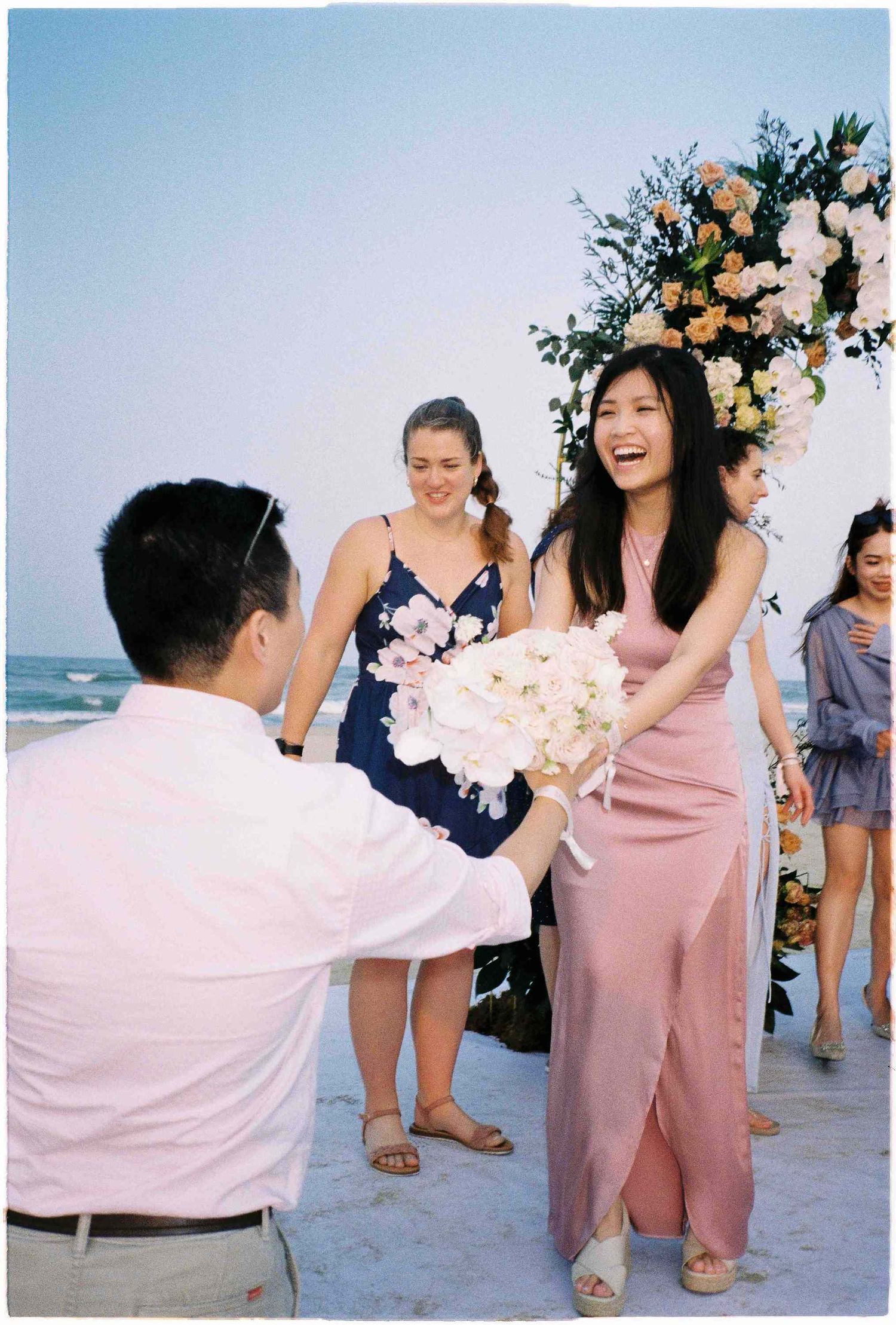 Woman in pink dress laughing while receiving bouquet at beach wedding ceremony with floral arch backdrop.